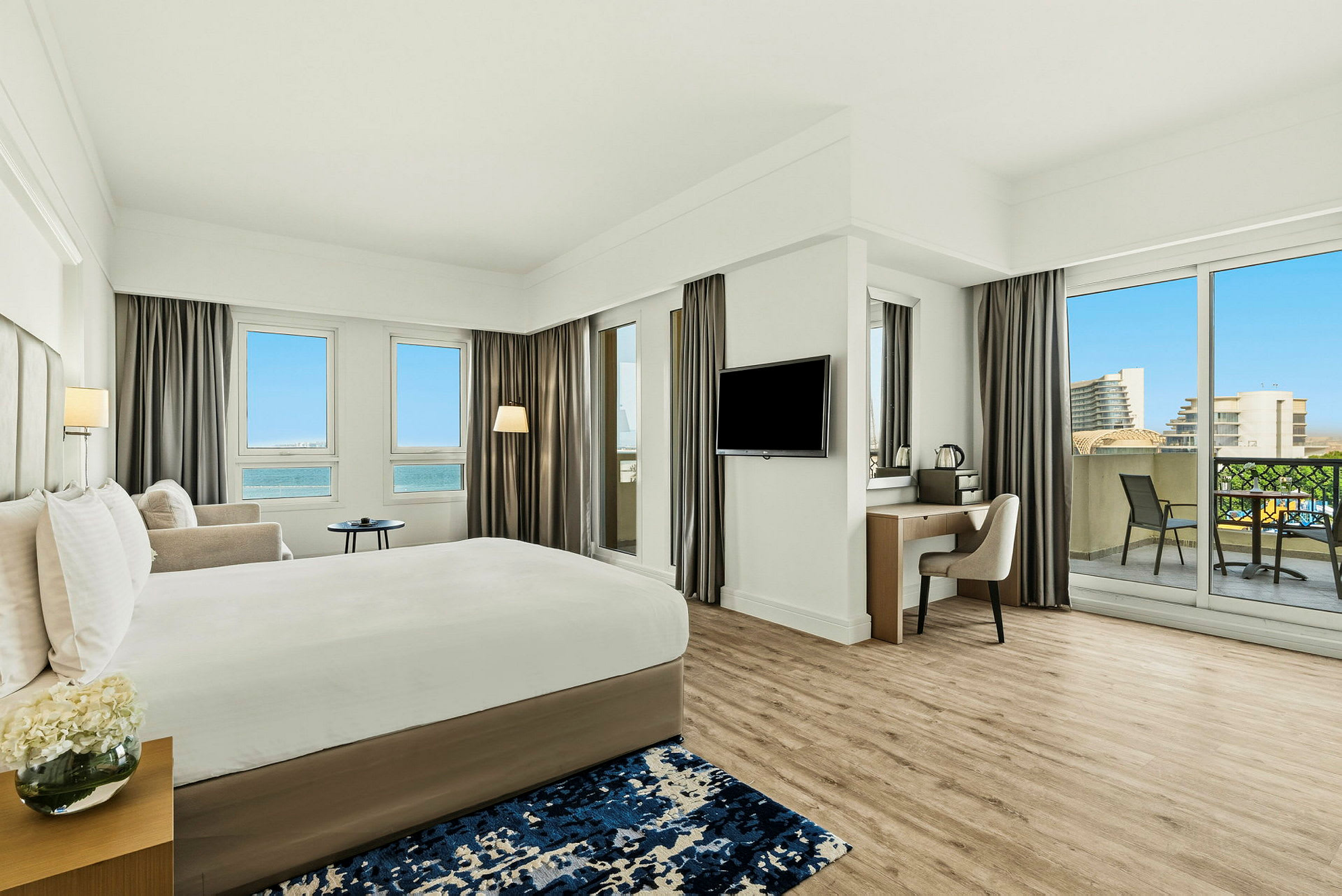 Et moderne hotellrom med en stor seng, skrivebord, balkong og utsikt over en blå himmel og bygninger. (AI-generert tekst)
