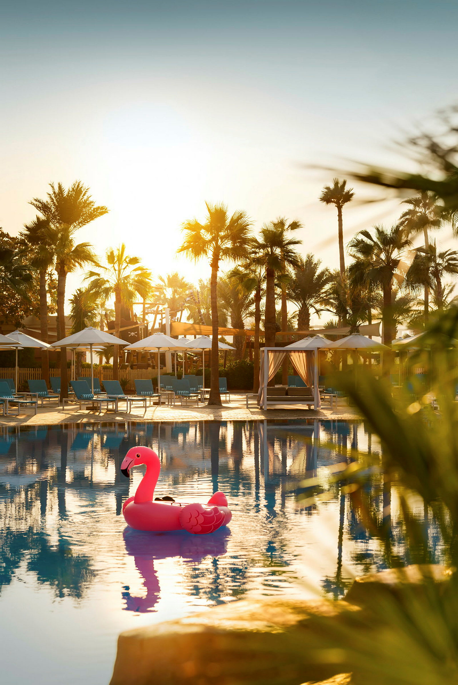 Rosa oppblåsbar flamingo i hotellbasseng med palmer og solsenger i bakgrunnen. (AI-generert tekst)