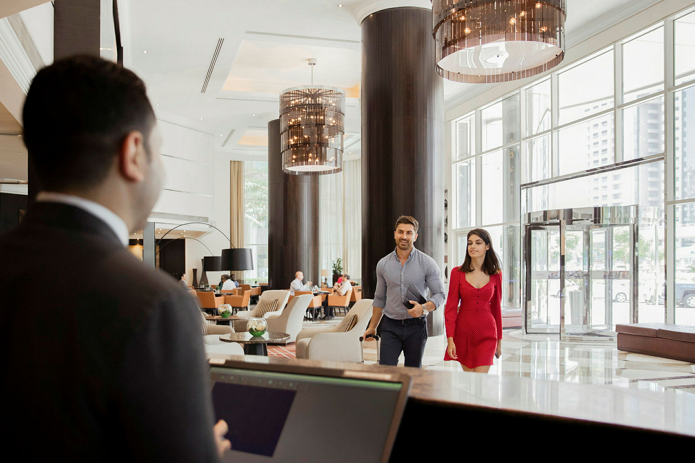 Interiør av en hotellobby med en resepsjon i forgrunnen og to gjester som går mot resepsjonen. (AI-generert tekst)