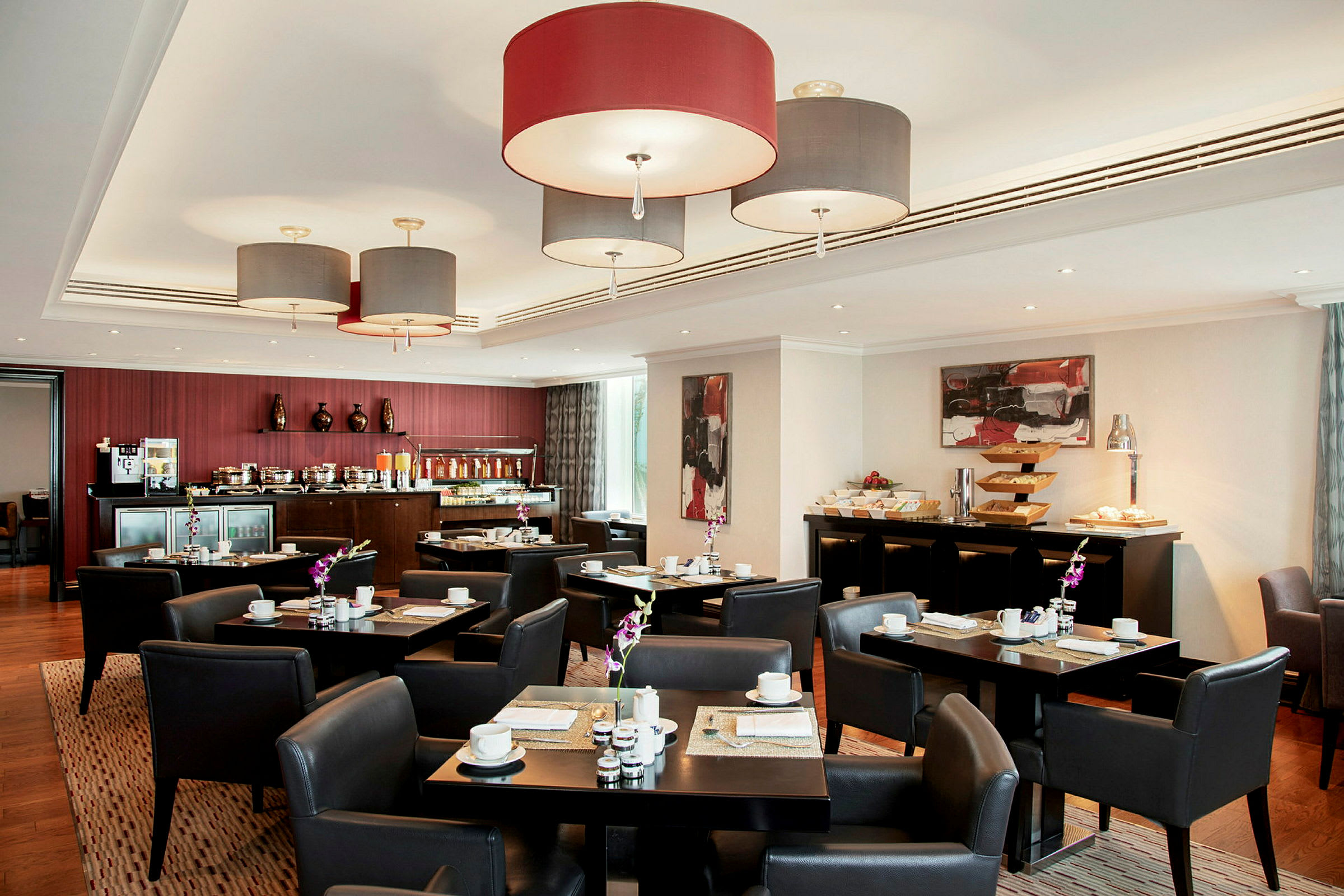 Et bistroområde med bord og stoler omgitt av en bufféservering. Hengende lamper og kunst på veggene kompletterer interiøret. (AI-generert tekst)
