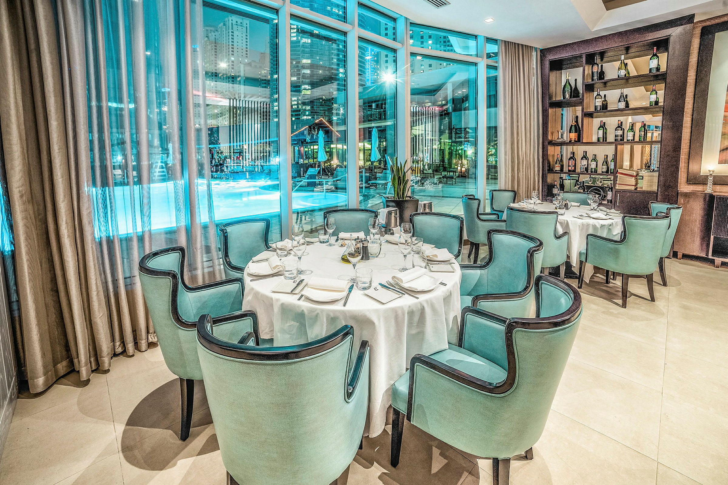 En elegant restaurant med dekkede bord og grønnblå stoler, plassert ved store vinduer som gir utsikt over et opplyst bassengområde om natten. (AI-generert tekst)