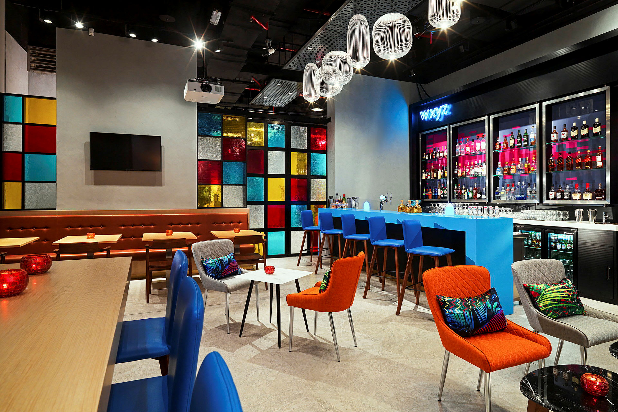 En moderne innredet bar eller lounge med fargerike møbler, en blå bardisk og hyller med flasker i bakgrunnen. Et vindspanel med ulike fargede ruter gir en livlig bakgrunn. Belysningen er varm, og designen er elegant og stilfull. (AI-generert tekst)