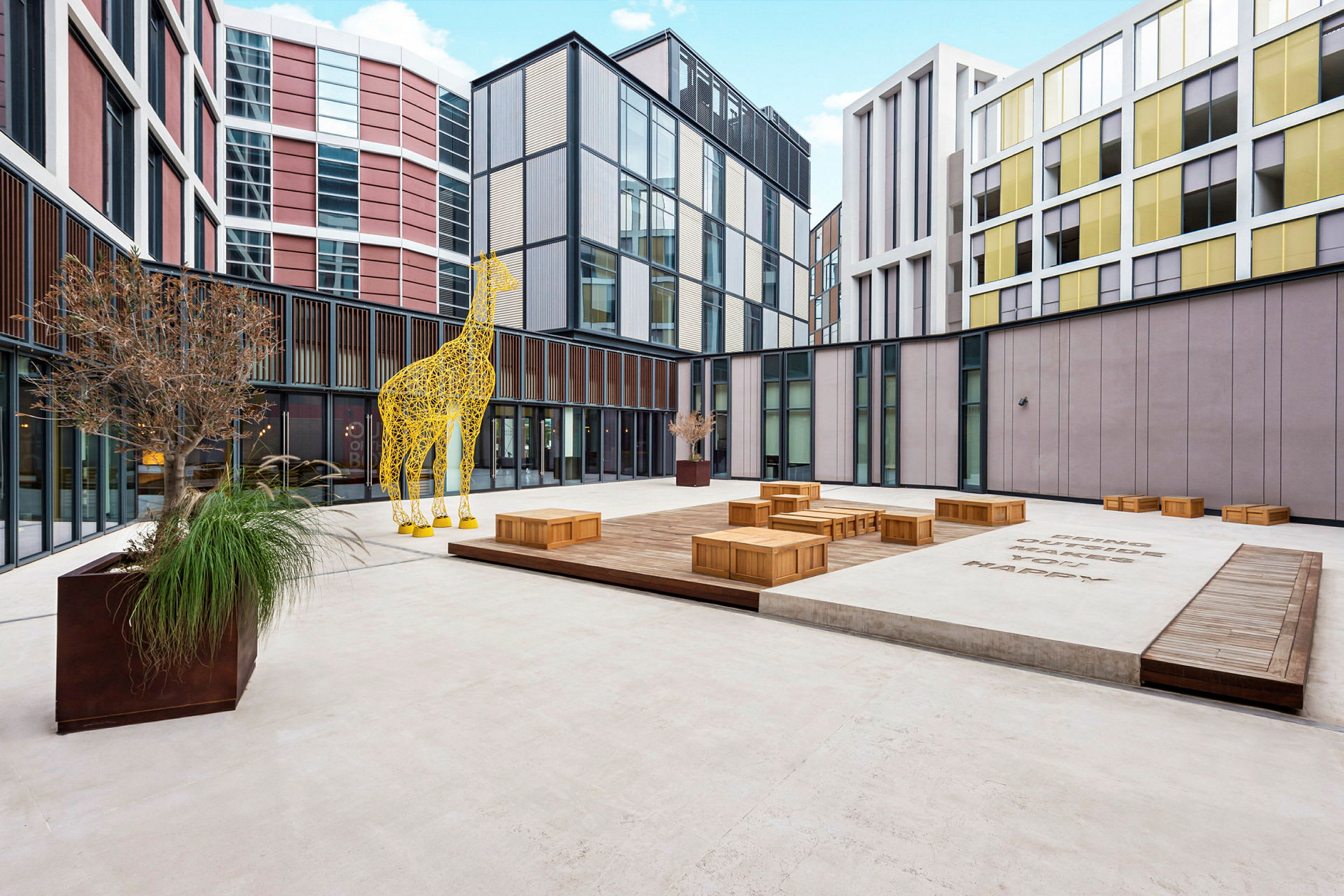 En indre gårdsplass med moderne bygninger, en gul giraffskulptur og trebokser. (AI-generert tekst)