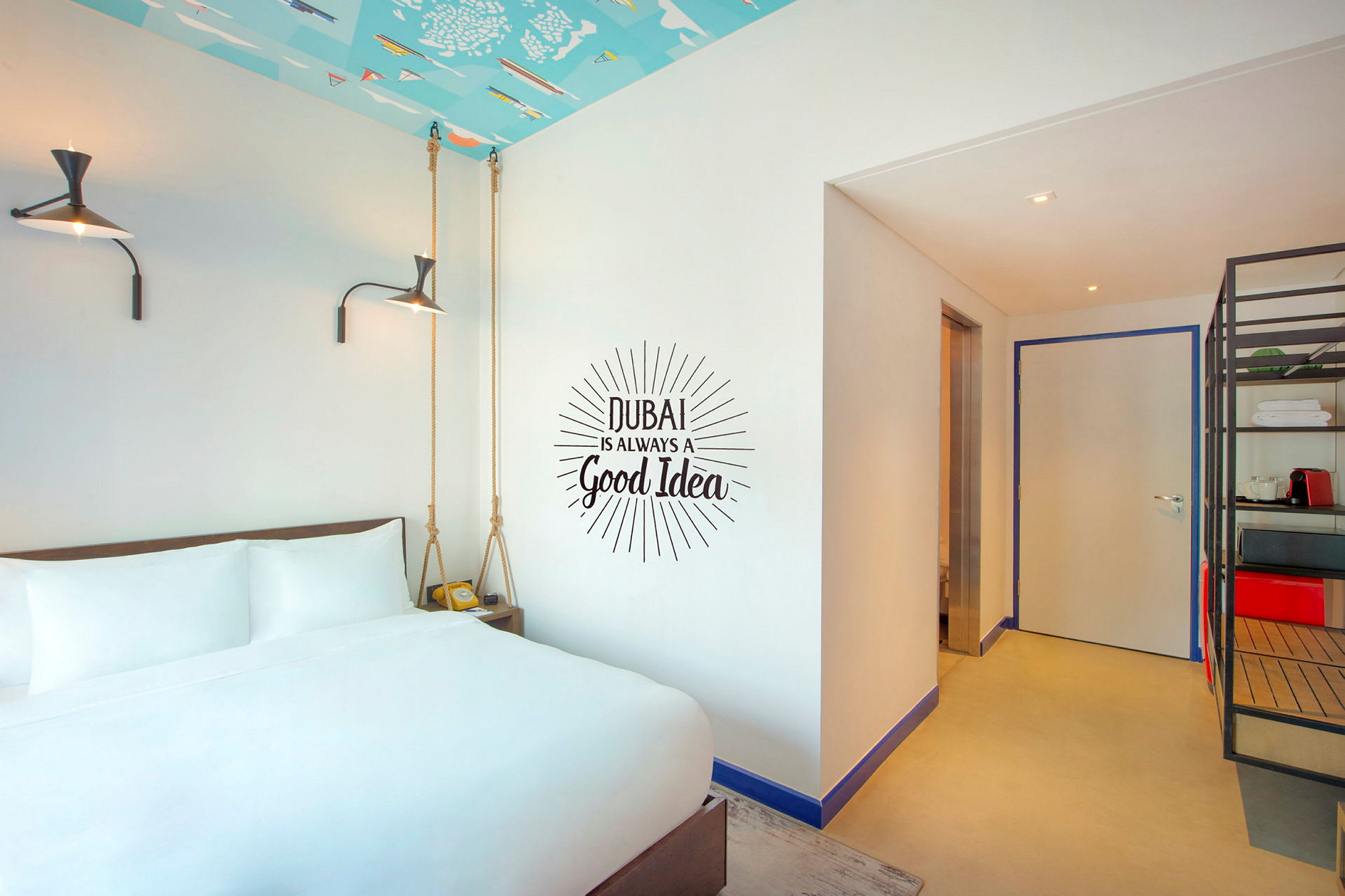 Hotellrom med hvit seng foran en vegg med teksten "Dubai is always a good idea". (AI-generert tekst)