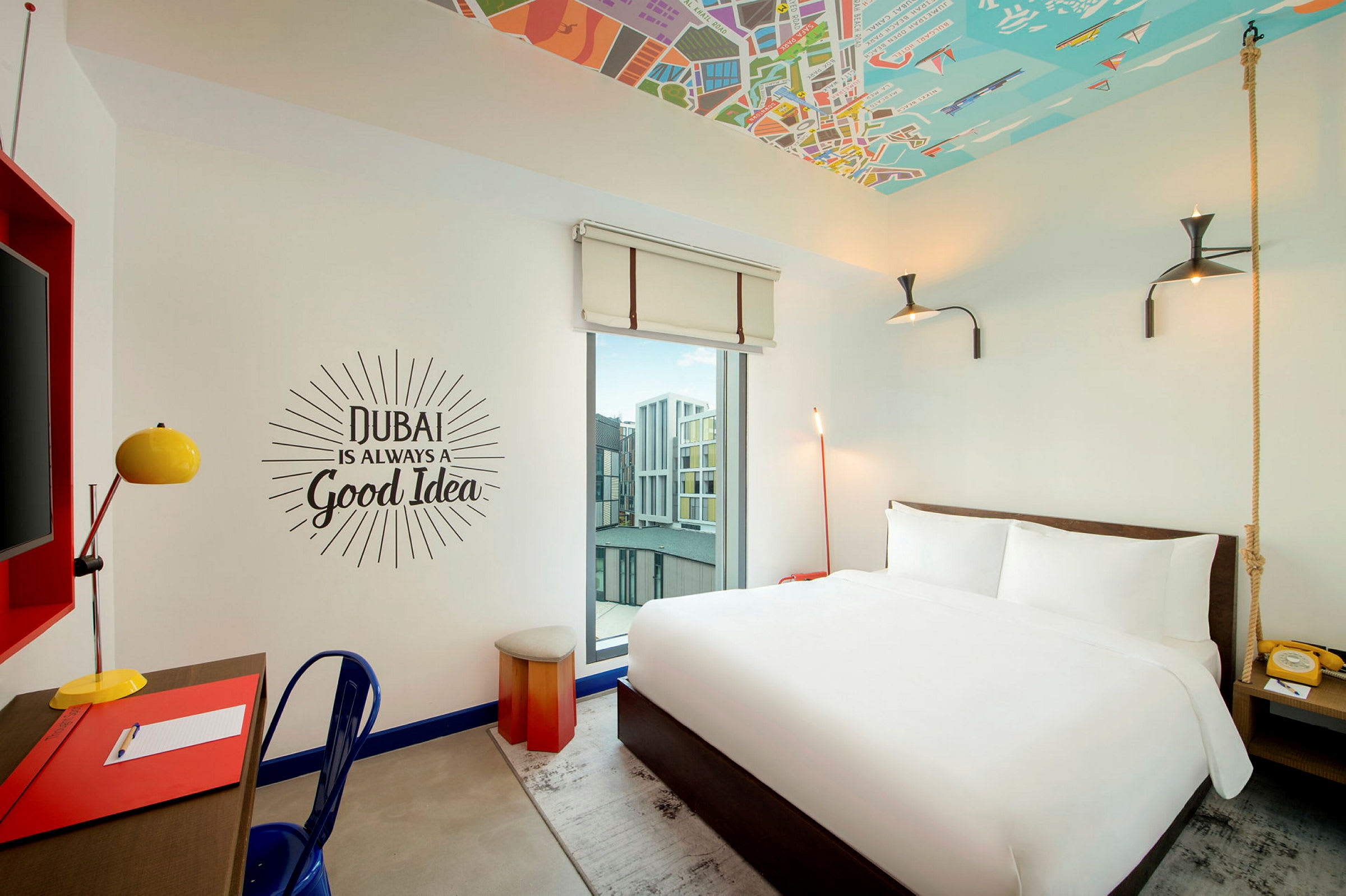 Et hotellrom med en hvit dobbeltseng, skrivebord med lampe og en veggtekst som sier 'Dubai is always a good idea'. (AI-generert tekst)