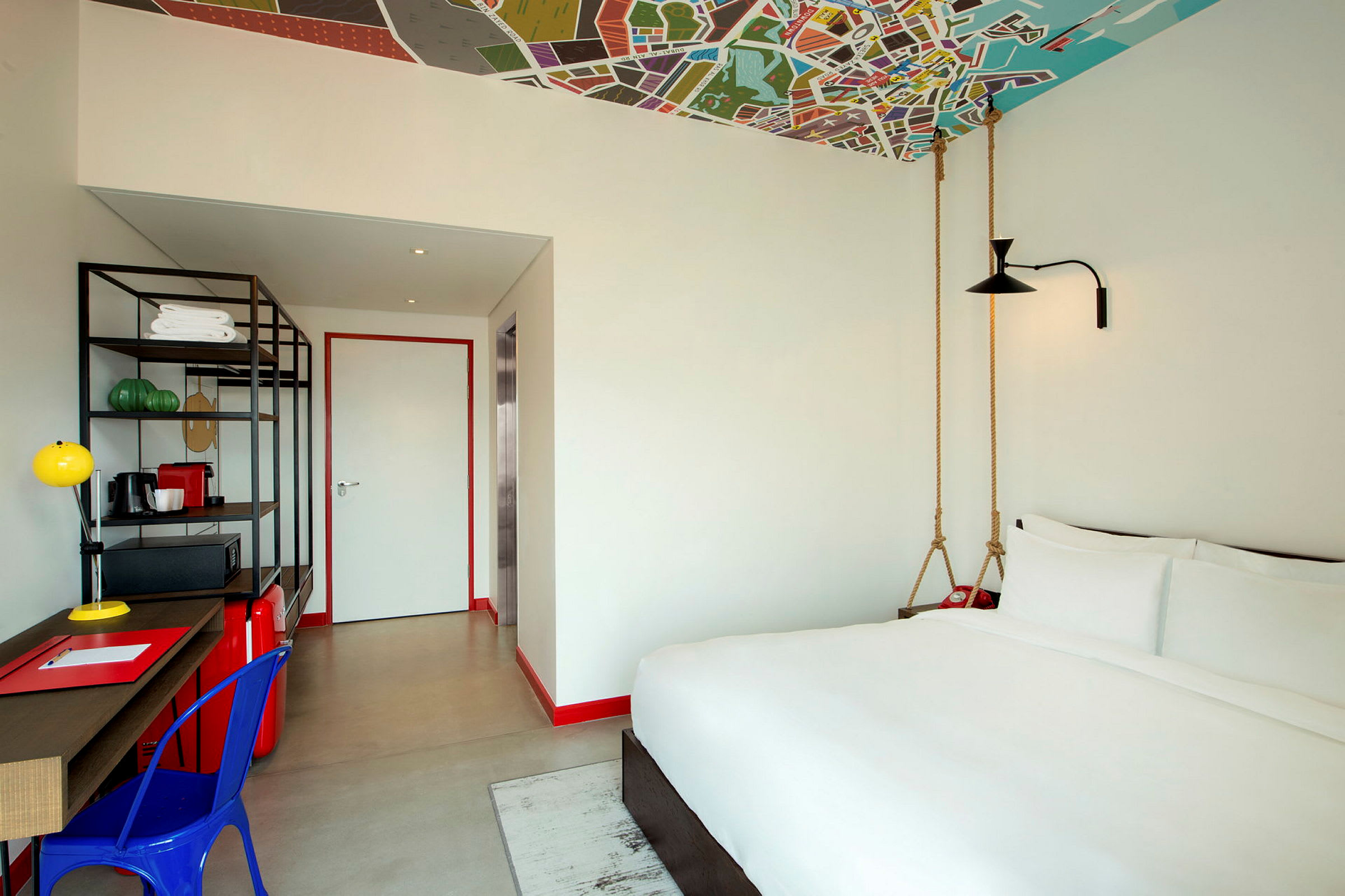 Et hotellrom med en seng, et skrivebord og en åpen hylle. Taket har et fargerikt design. (AI-generert tekst)