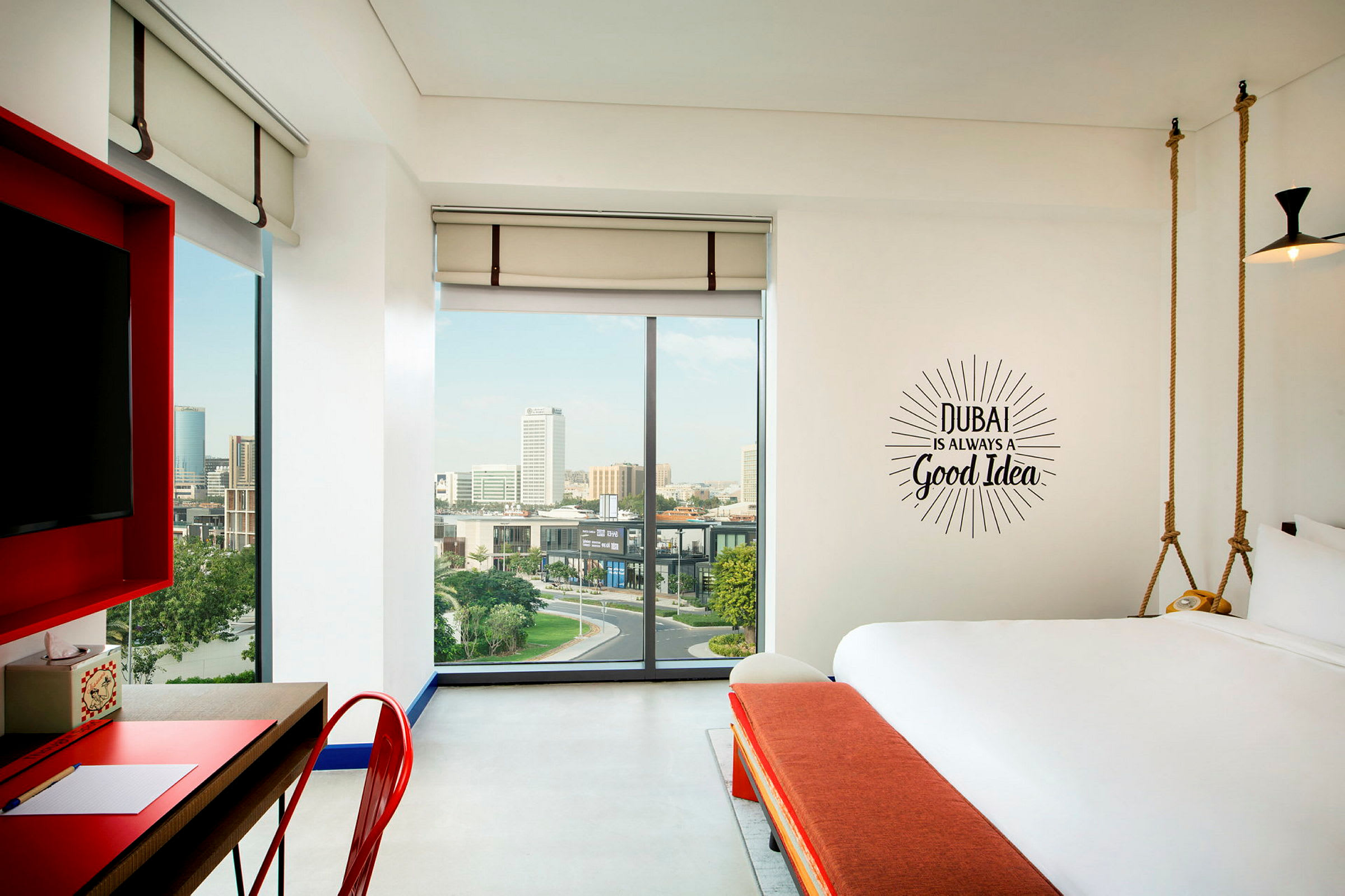 Et moderne hotellrom med store vinduer og utsikt over en by. På veggen står teksten: "Dubai is always a good idea". (AI-generert tekst)