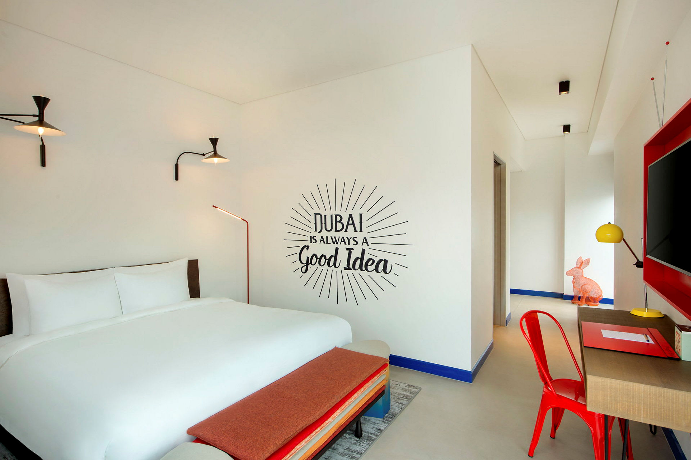Et hotellrom med en dobbeltseng, en veggtekst som lyder "Dubai is always a good idea", og innredning i moderne farger. (AI-generert tekst)