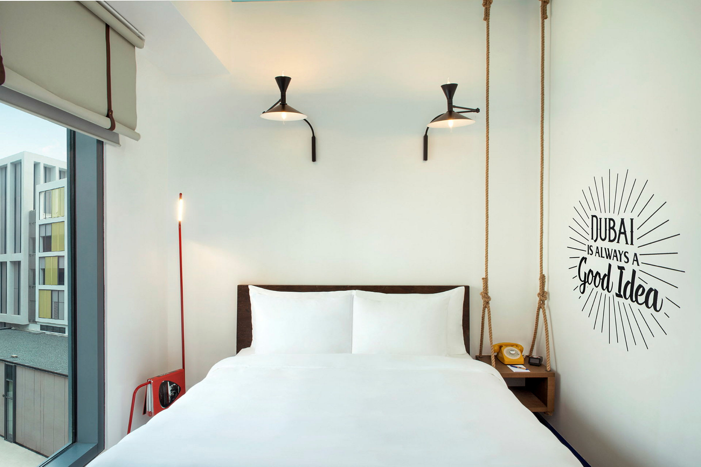 Et moderne hotellrom med en seng, to lamper på veggen og en veggtekst som lyder "Dubai is always a good idea". (AI-generert tekst)