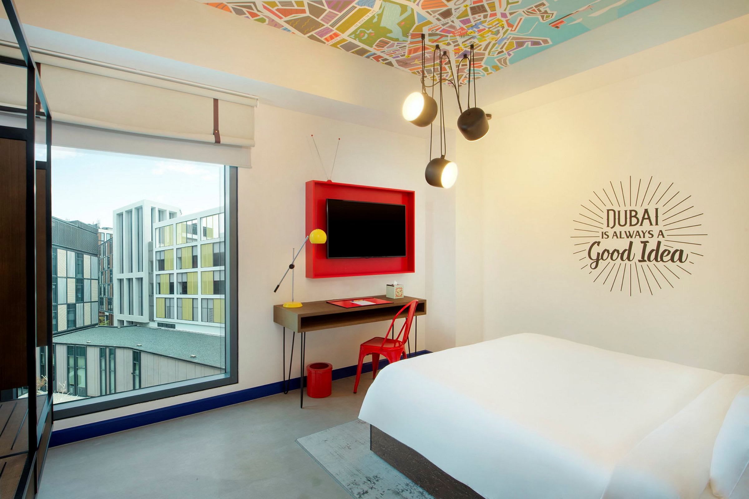 Et lyst hotellrom med en stor seng, skrivebord og en vegg-TV. Vinduet viser bygninger utenfor. (AI-generert tekst)