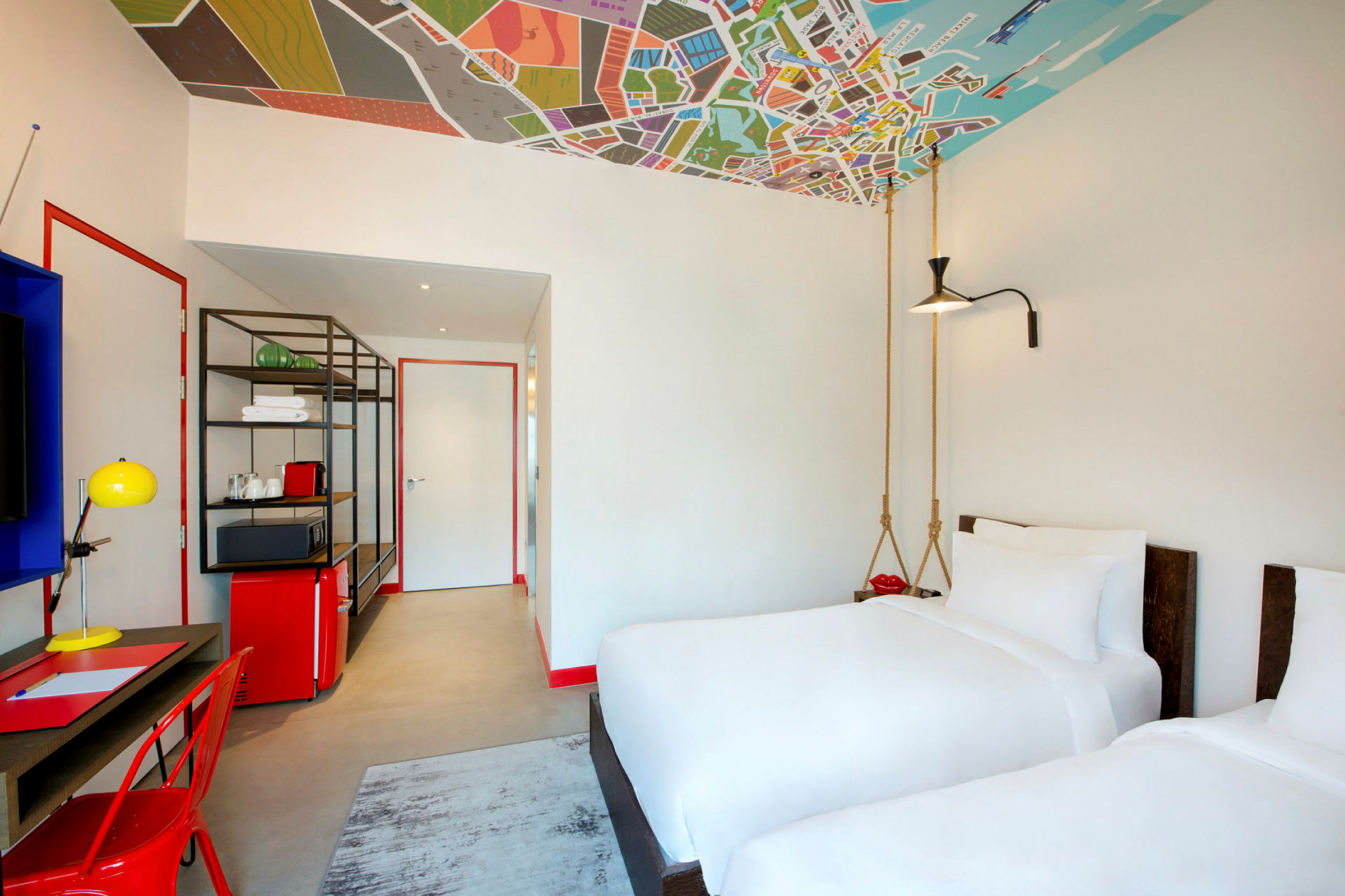 Et moderne hotellrom med to hvite enkeltsenger, et skrivebord, en rød stol og fargerike interiørdetaljer. Taket er dekorert med et kartmønster. (AI-generert tekst)