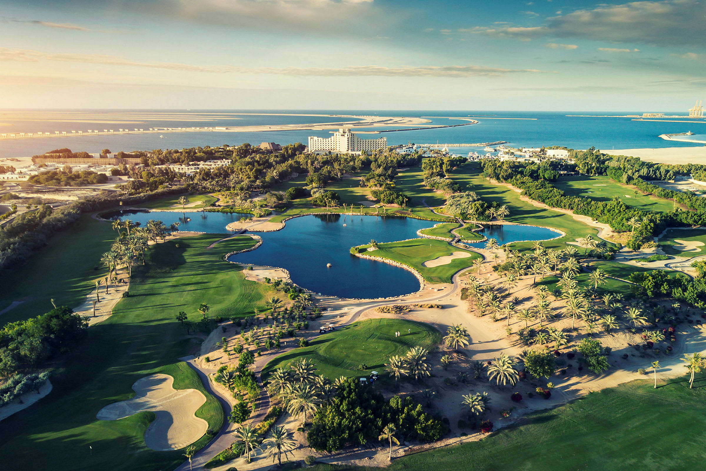 Flyfoto av en golfbane med vannhindringer og omkringliggende vegetasjon, samt hav og bygninger i bakgrunnen. (AI-generert tekst)