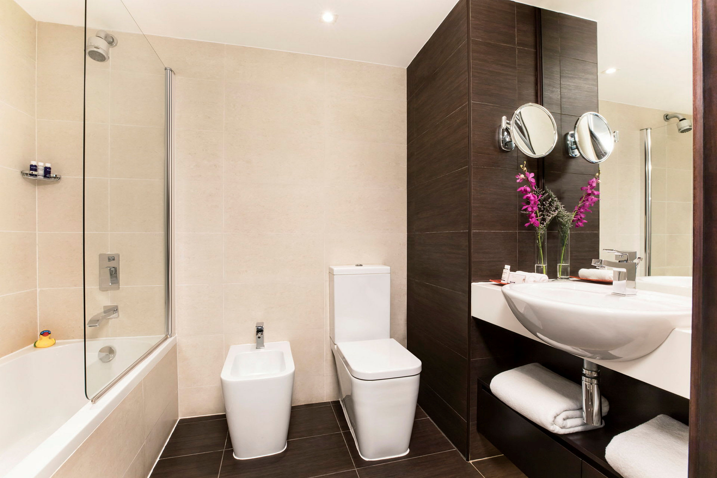 Et moderne bad med et badekar, et toalett, et bidet og en servant med speil. Dekorasjonen inkluderer rosa blomster i glassvaser. (AI-generert tekst)