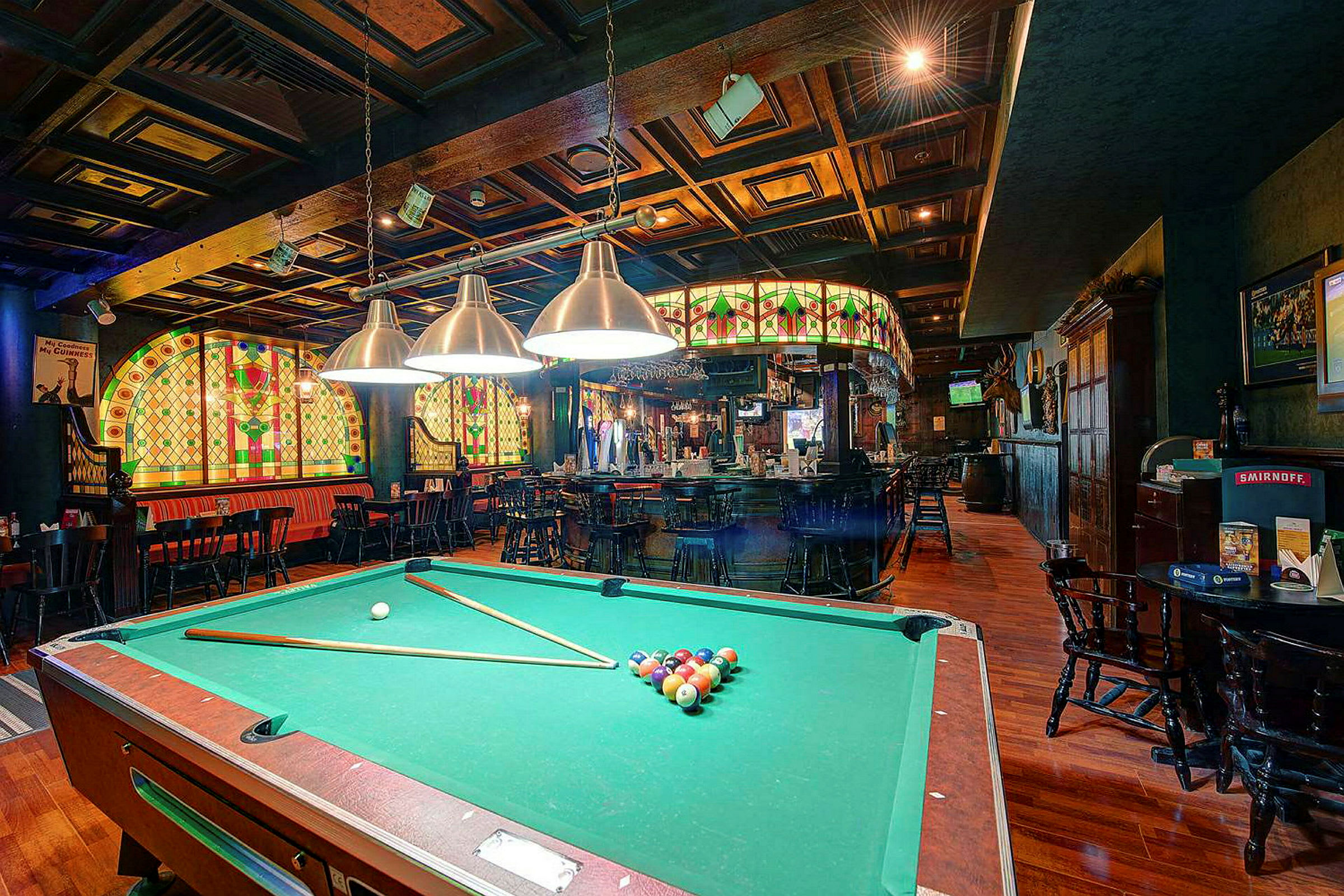 Et biljardbord står i sentrum av en koselig pub, omgitt av belysning og dekorerte glassmalerier. (AI-generert tekst)