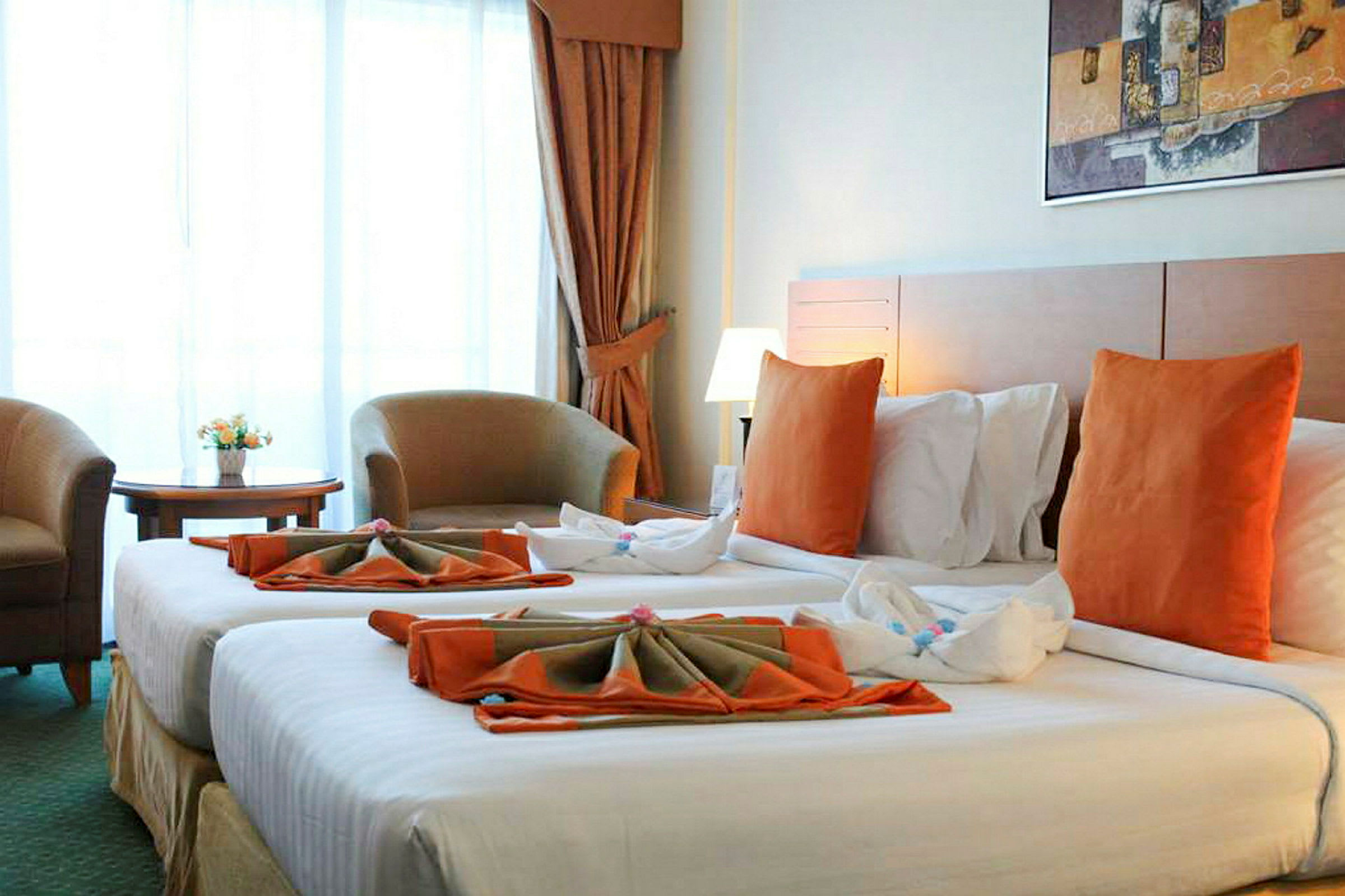 Et hotellrom med to senger oppredd med hvitt sengetøy og oransje puter. Det er en sittegruppe med to lenestoler og et lite bord foran et stort vindu, samt et maleri på veggen over sengene. (AI-generert tekst)