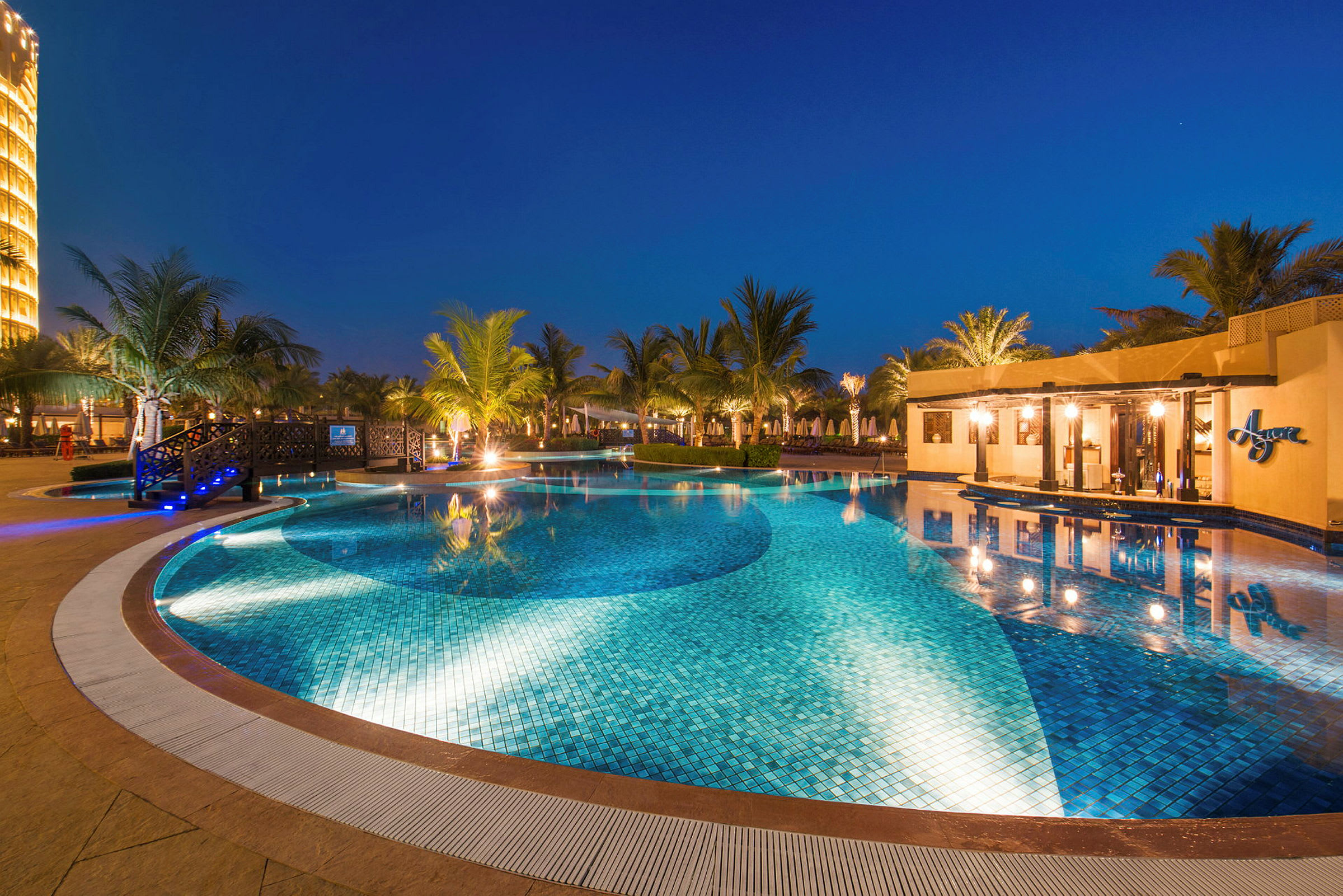 Et utendørs svømmebasseng omgitt av palmetrær og bygninger, opplyst om kvelden. (AI-generert tekst)