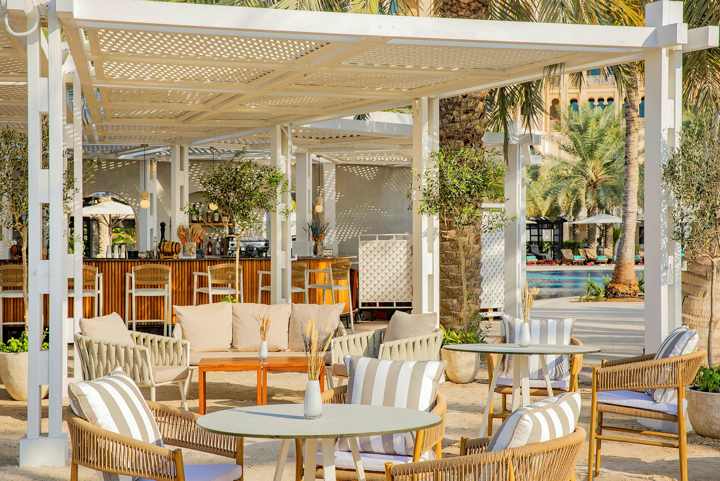 Utendørs loungeområde med sittemøbler og bord under en hvit pergola, omgitt av palmetrær og grøntområder. (AI-generert tekst)