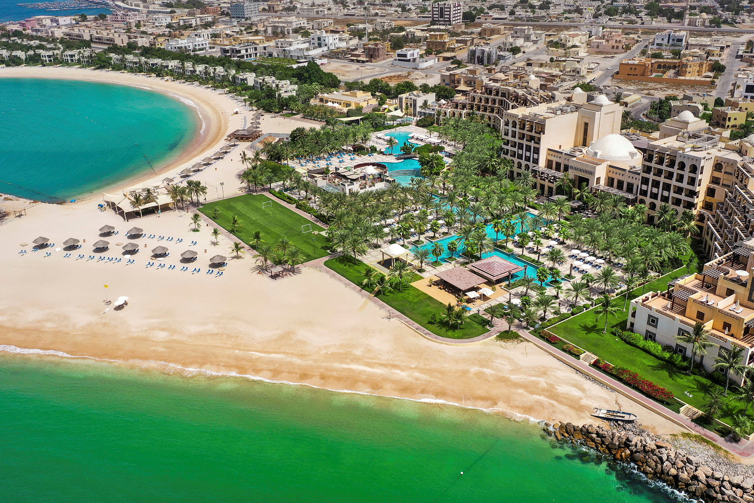 Luftfoto av et hotellresort ved stranden med bassenger, gressmatter og palmer. Havet og sandstranden er også synlige. (AI-generert tekst)