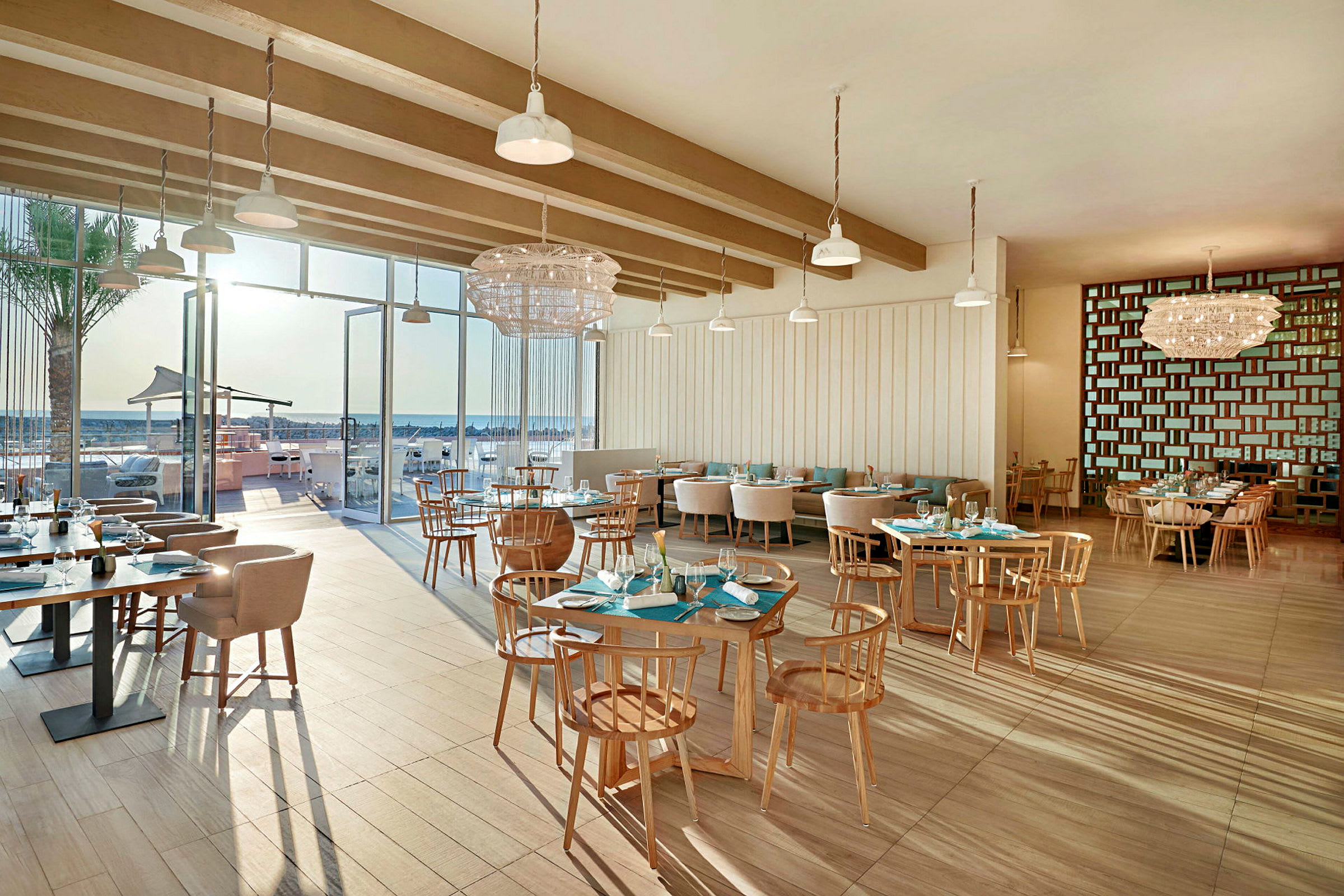 En moderne restaurant innendørs med store glassvinduer vendt mot en uteservering og stranden. (AI-generert tekst)
