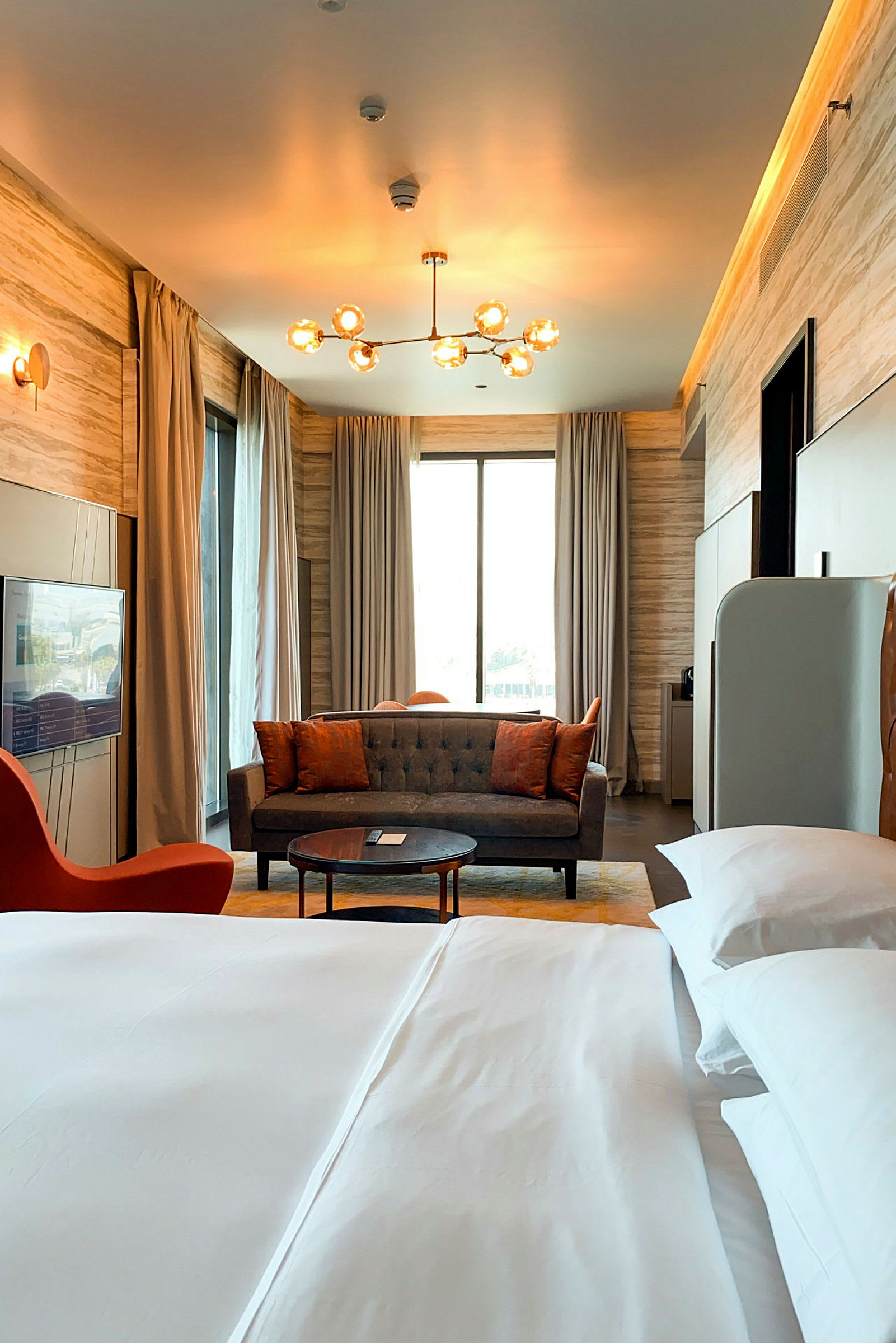 Et elegant møblert hotellrom med en stor seng i forgrunnen og en sofagruppe foran et stort vindu med gardiner. En moderne lampe henger i taket. (AI-generert tekst)