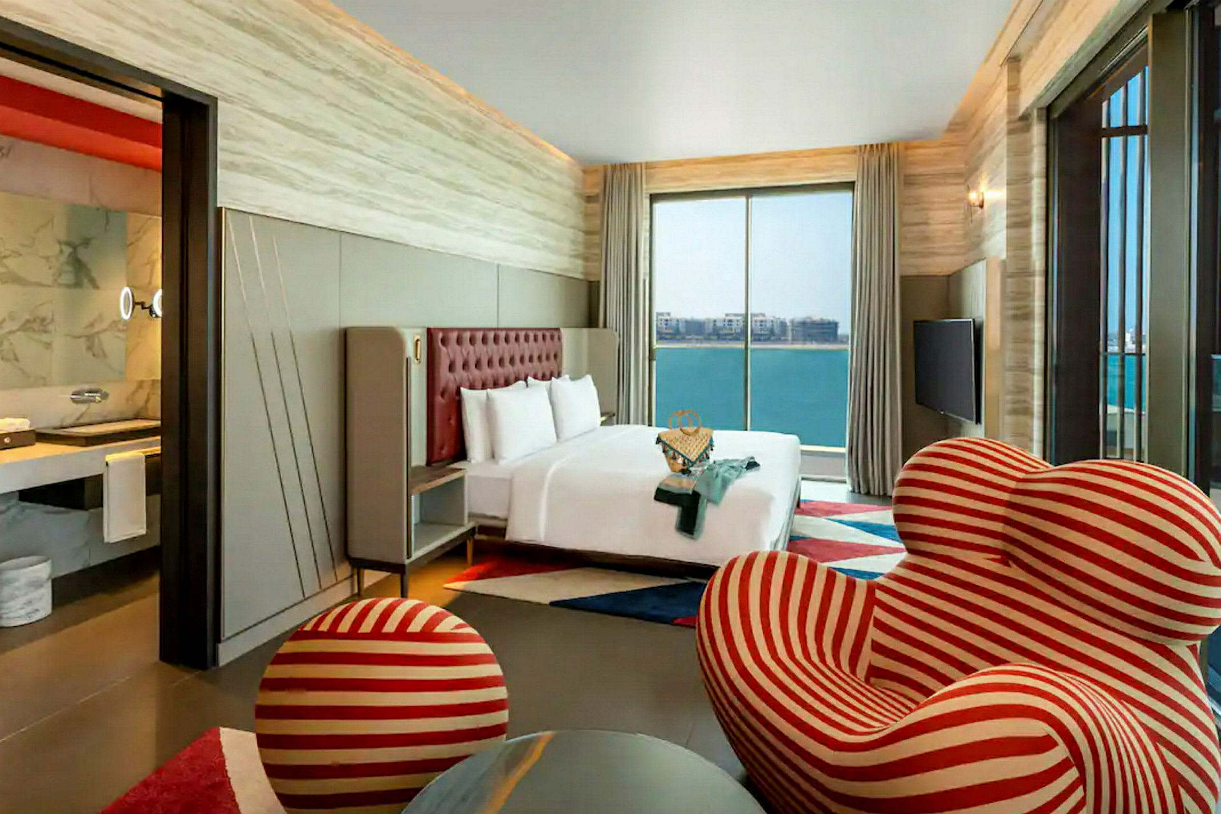 Et moderne hotellrom med en stor seng foran et stort vindu som viser utsikt over havet. Rommet har også en unik stripete rød og hvit lenestol med en matchende fotskammel. (AI-generert tekst)