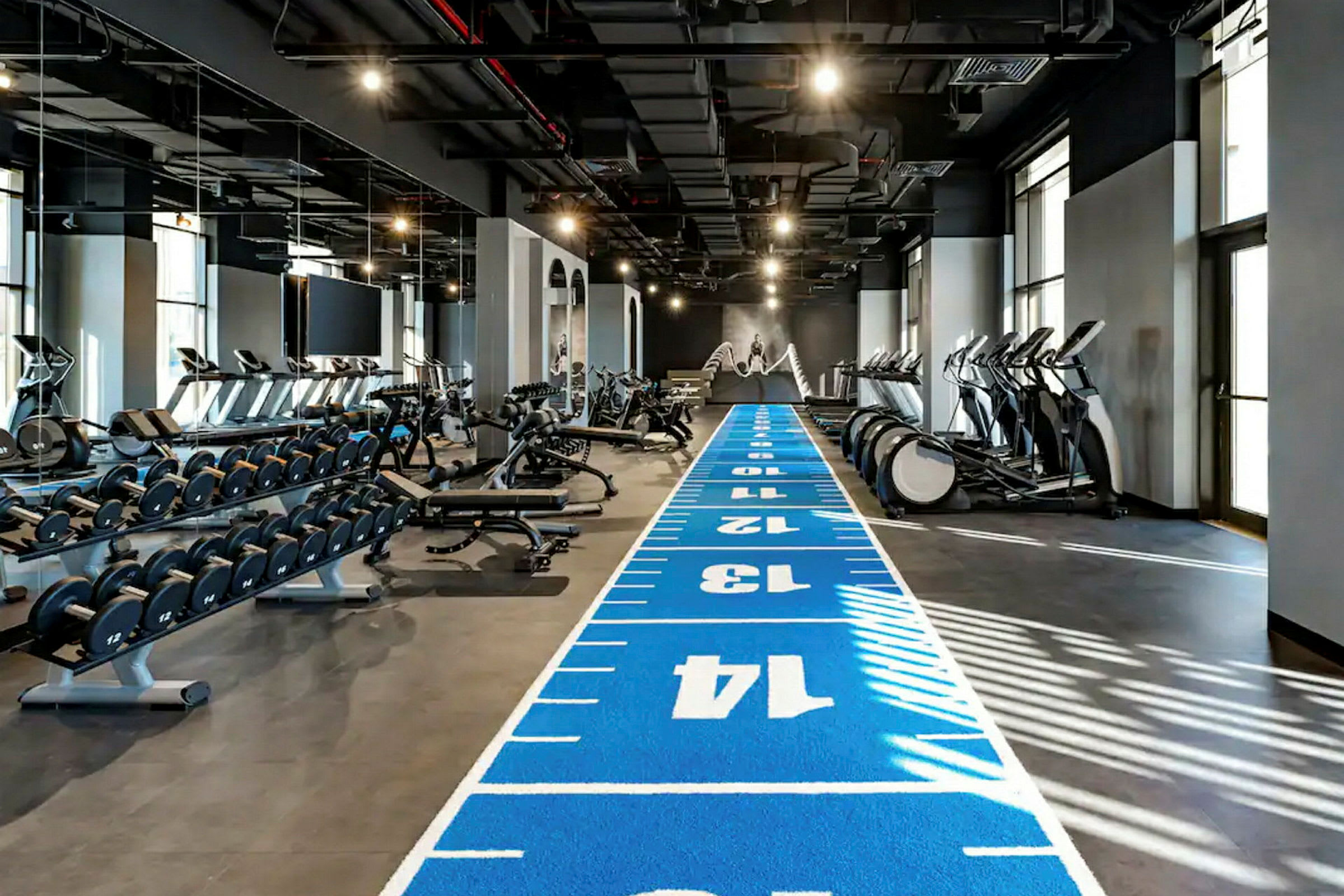 Et treningsstudio med treningsutstyr, vekter og en blå løpematte merket med tall. (AI-generert tekst)