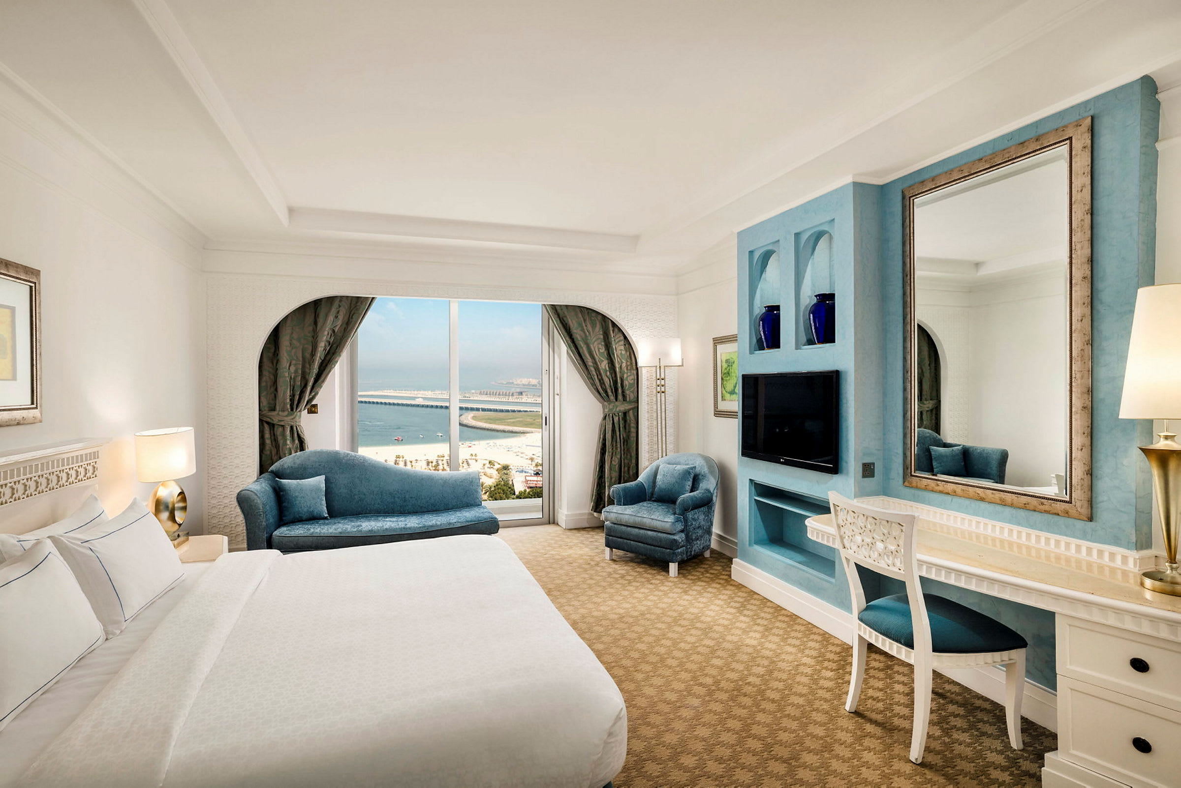Hotellrom med seng, sofaer, skrivebord og speil. Utsikt mot havet gjennom vinduet. (AI-generert tekst)