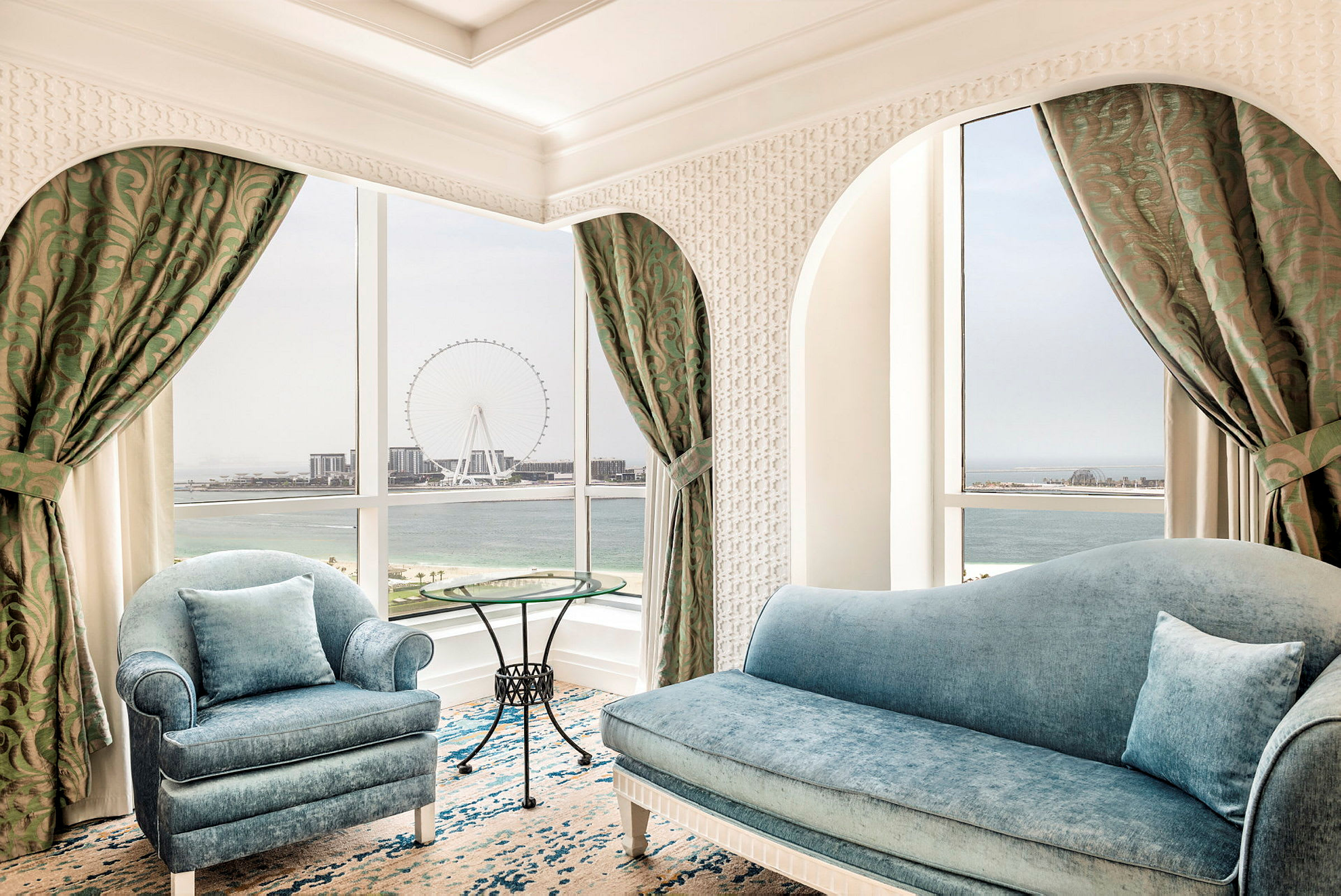 Et elegant rom med blå sofaer og et rundt glassbord. Vinduet har utsikt over havet og et stort pariserhjul. (AI-generert tekst)