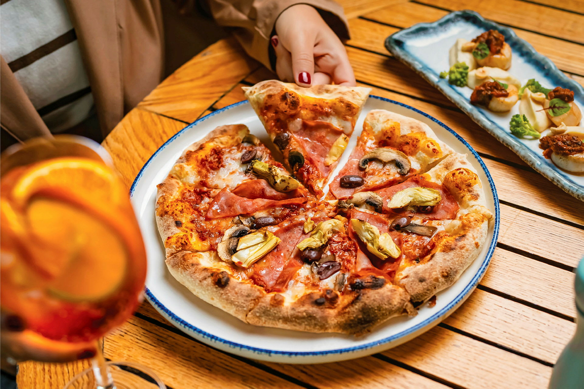 En tallerken med pizza toppet med skinke, oliven, sopp og artisjokker på et bord. En person tar et pizzastykke. Bak pizzaen er det et fat med små forretter og ved siden av et glass med oransje drikk. (AI-generert tekst)
