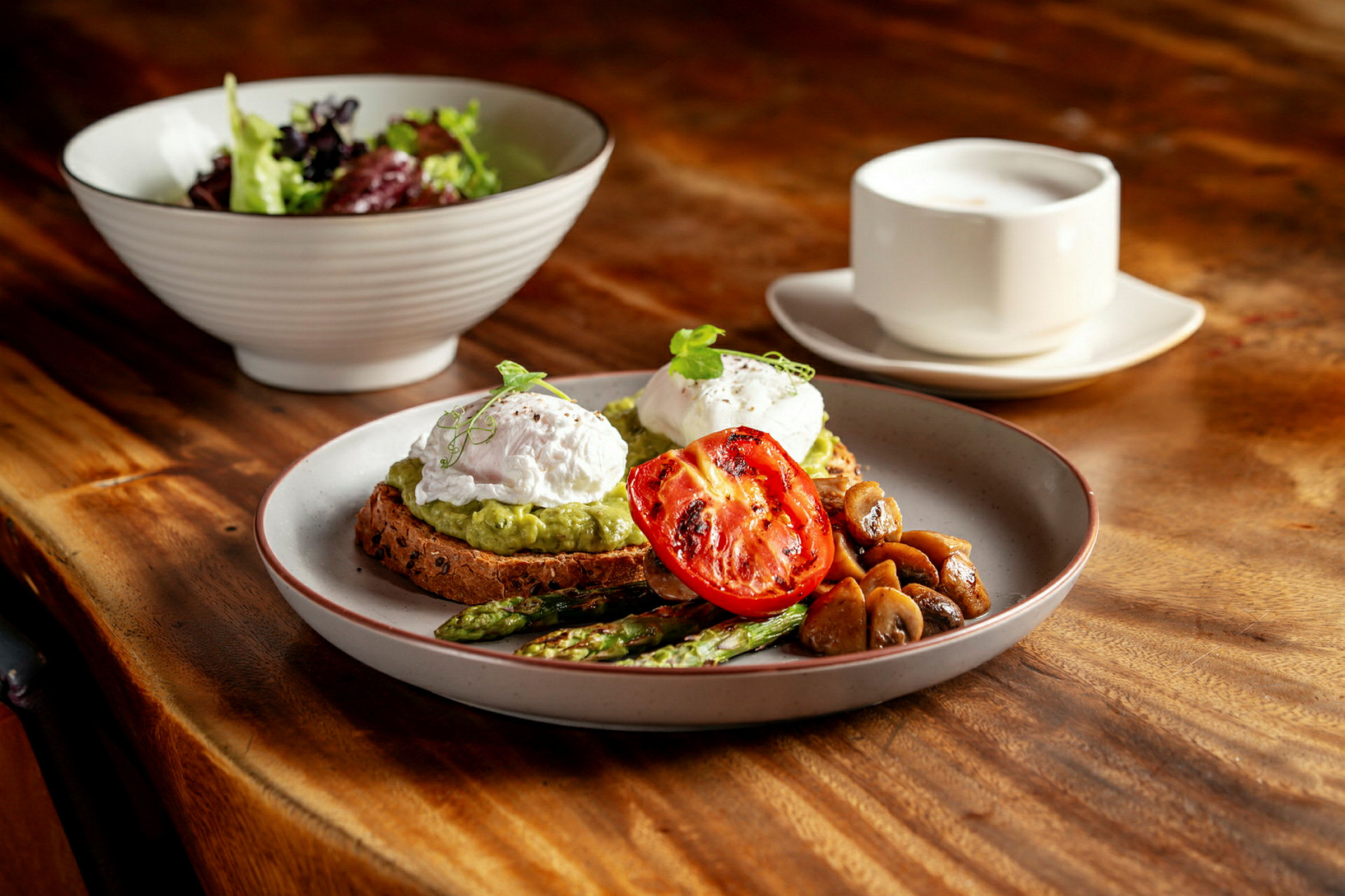 En tallerken med toast, most avokado, posjerte egg, grillet tomat, asparges og sopp. Bak ses en salatbolle og en kopp på et trebord. (AI-generert tekst)