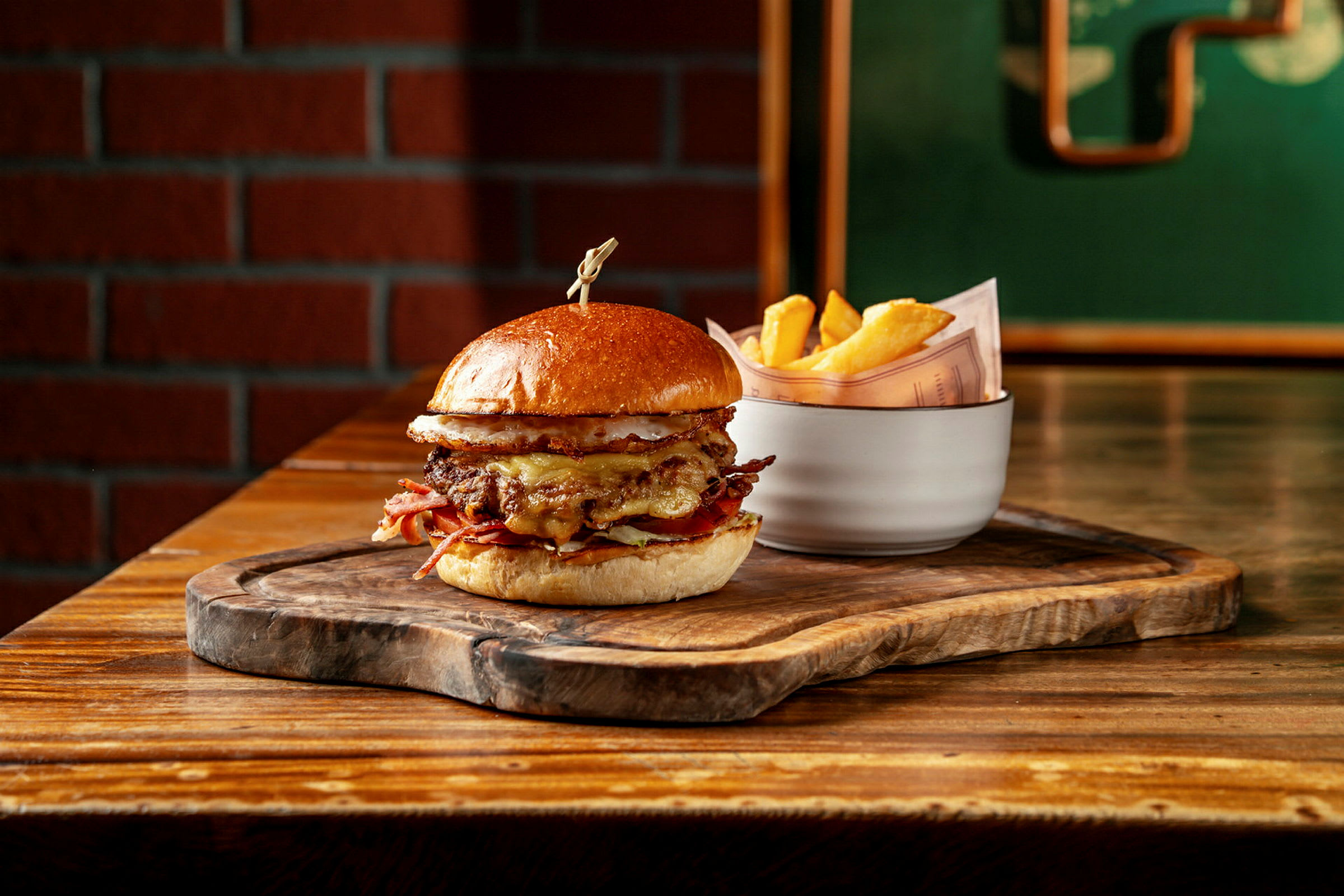 En hamburger med brød, kjøtt og ost servert på et trebrett. Ved siden av står en hvit skål med pommes frites. (AI-generert tekst)