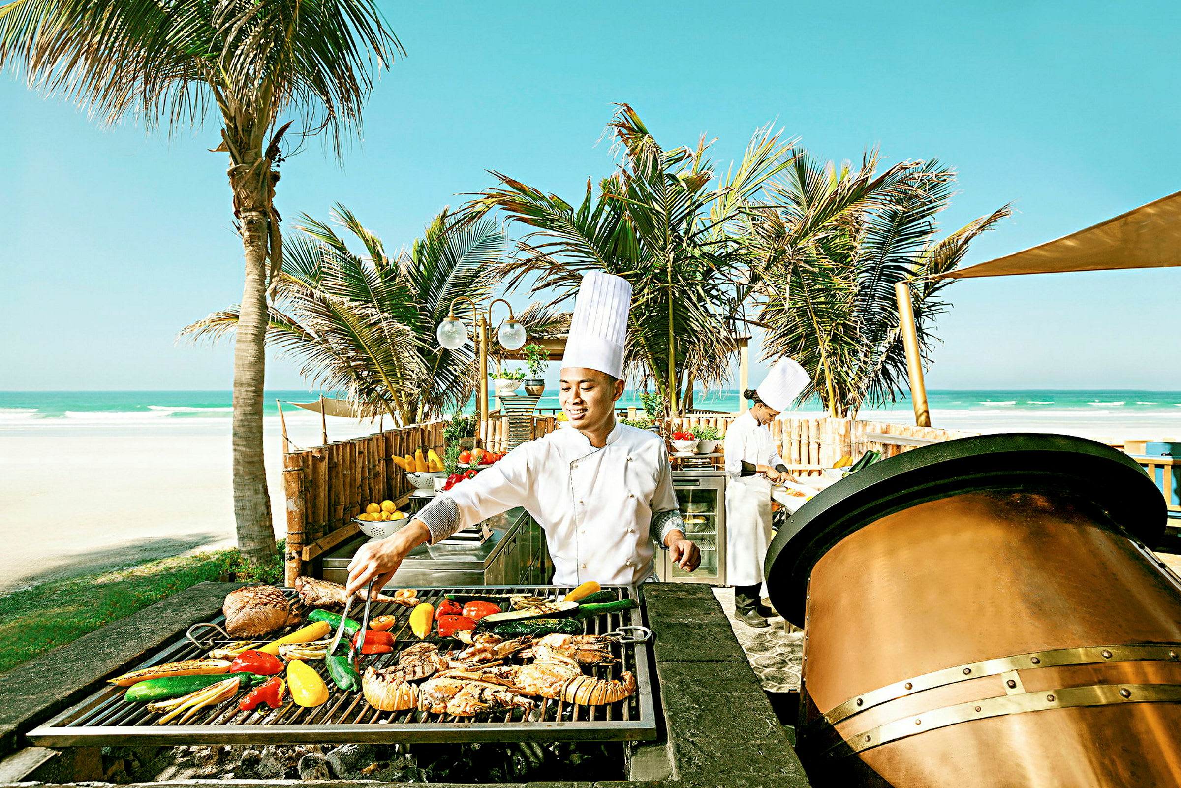 En kokk ved en grillstasjon ved stranden med palmer i bakgrunnen. (AI-generert tekst)