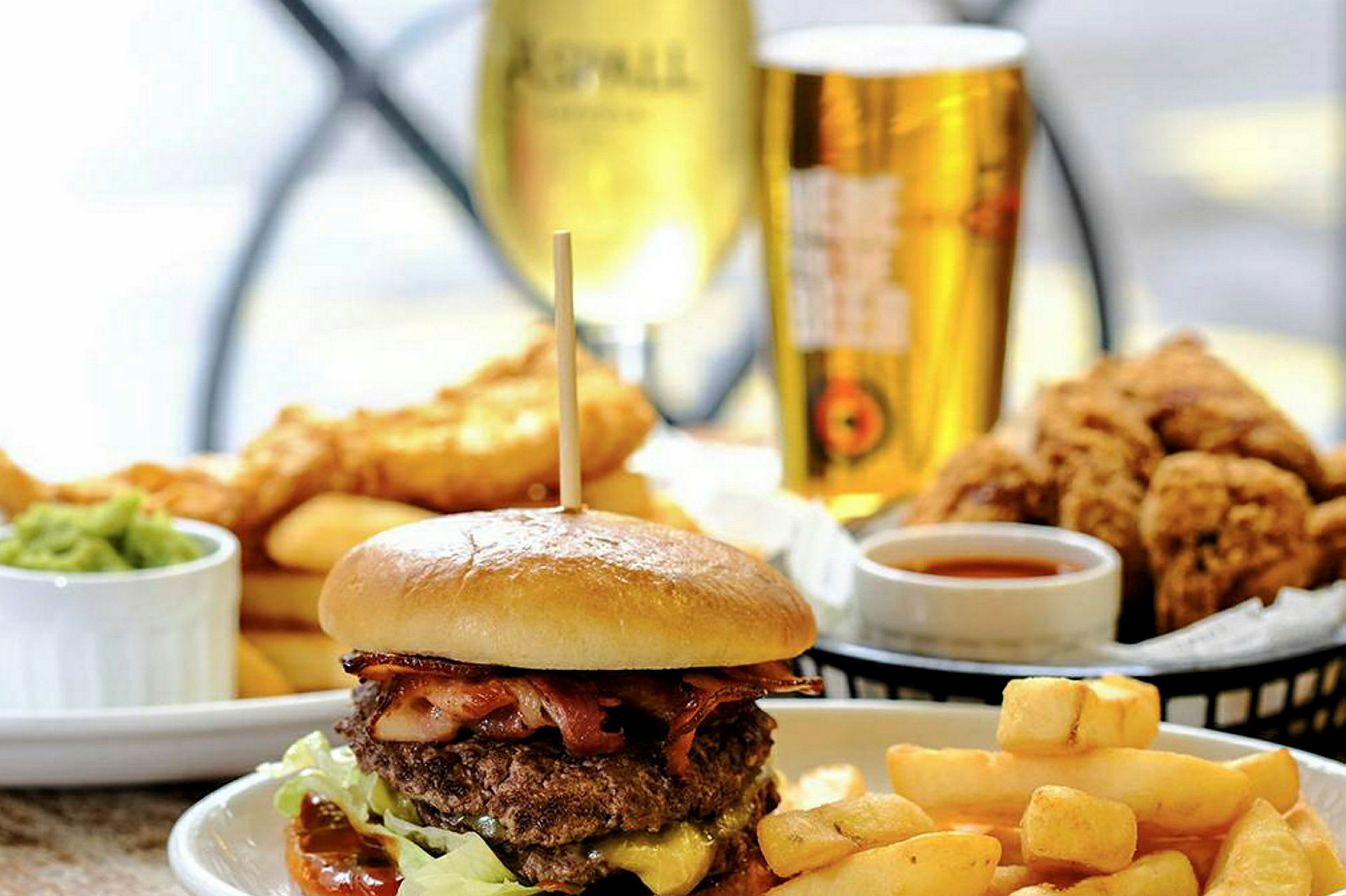 En kjøttburger med pommes frites og tilbehør, drikke og fritert mat i bakgrunnen. (AI-generert tekst)