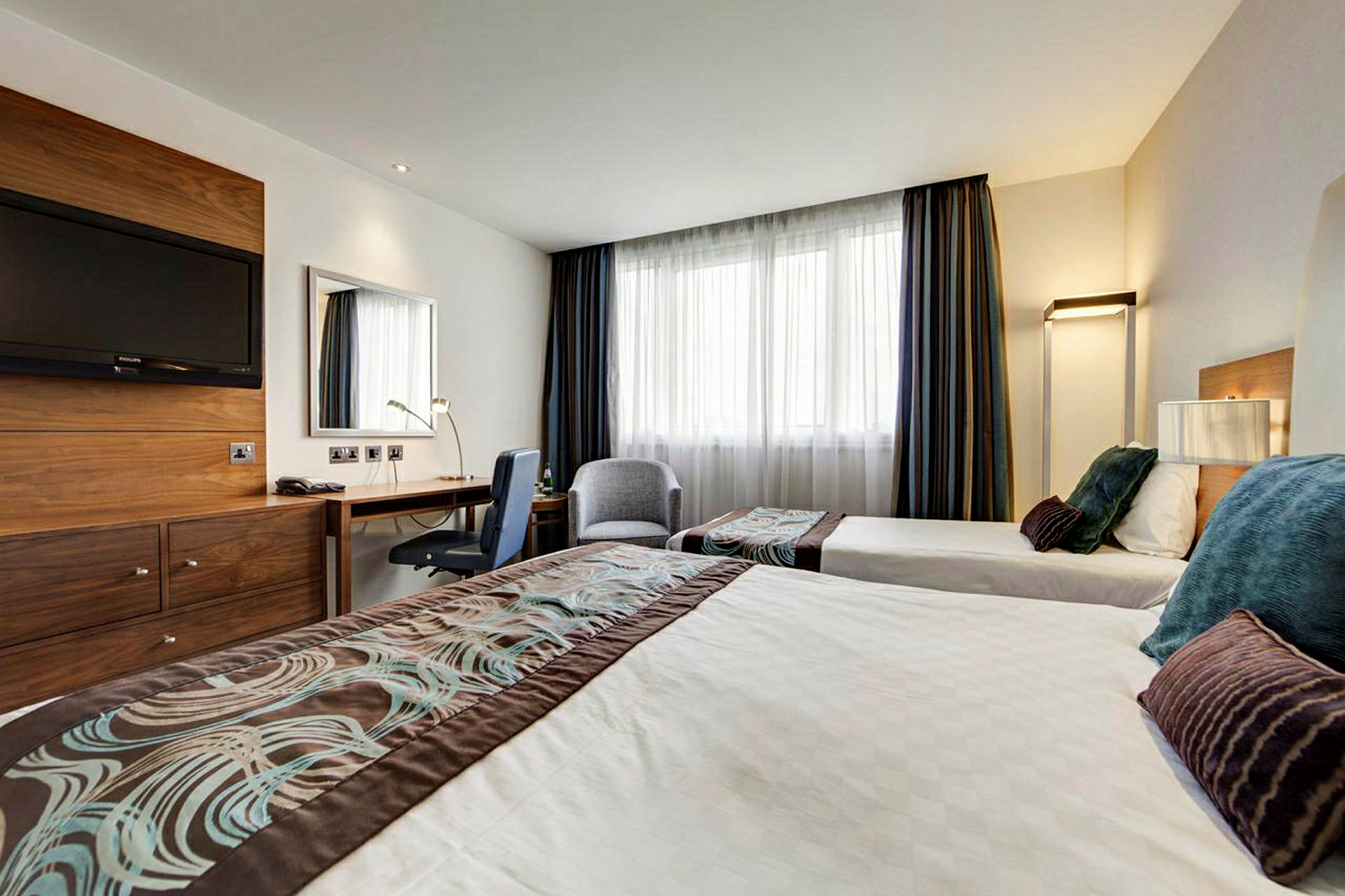 Et hotellrom med to enkeltsenger, et skrivebord og en TV på veggen. (AI-generert tekst)