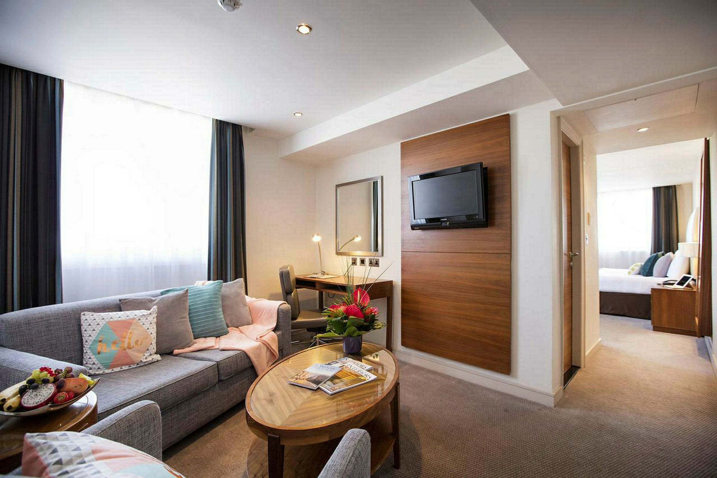 Et hotellrom med sittegruppe, sofabord, skrivebord, veggmontert TV og blomster. (AI-generert tekst)