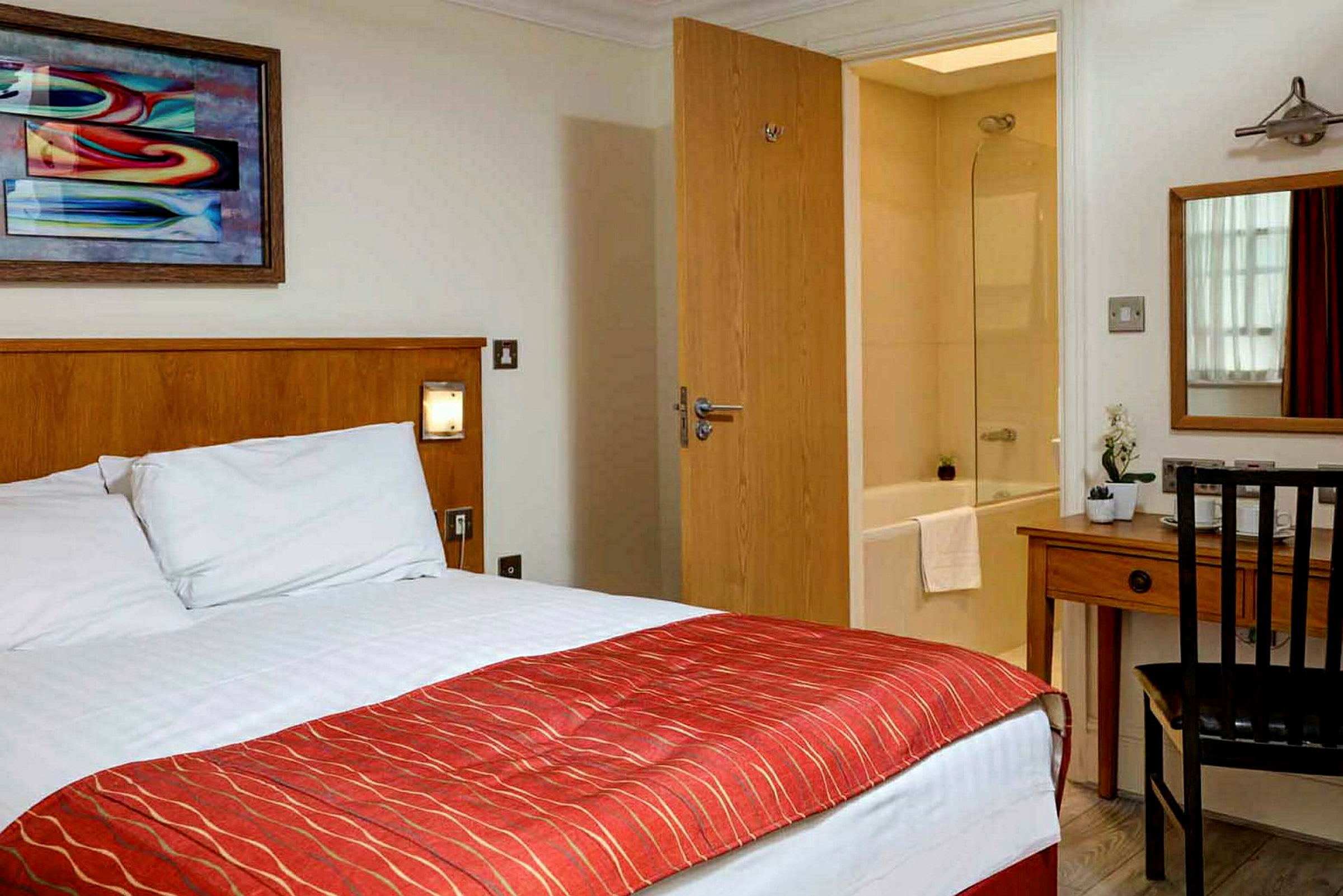 Hotellrom med seng, skrivebord, speil og utsikt mot et bad. (AI-generert tekst)