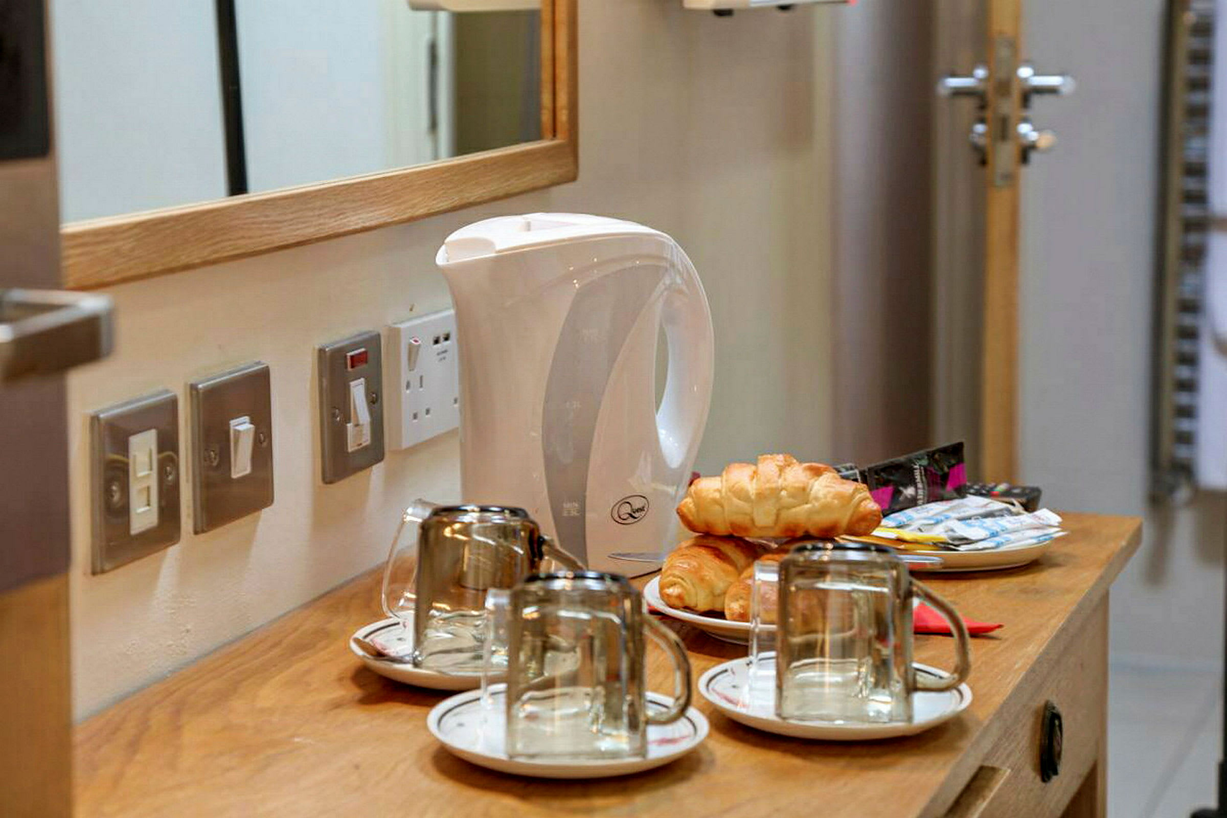 Vannkoker, kaffekopper med fat, croissanter og kaffeposer på en trebord. (AI-generert tekst)