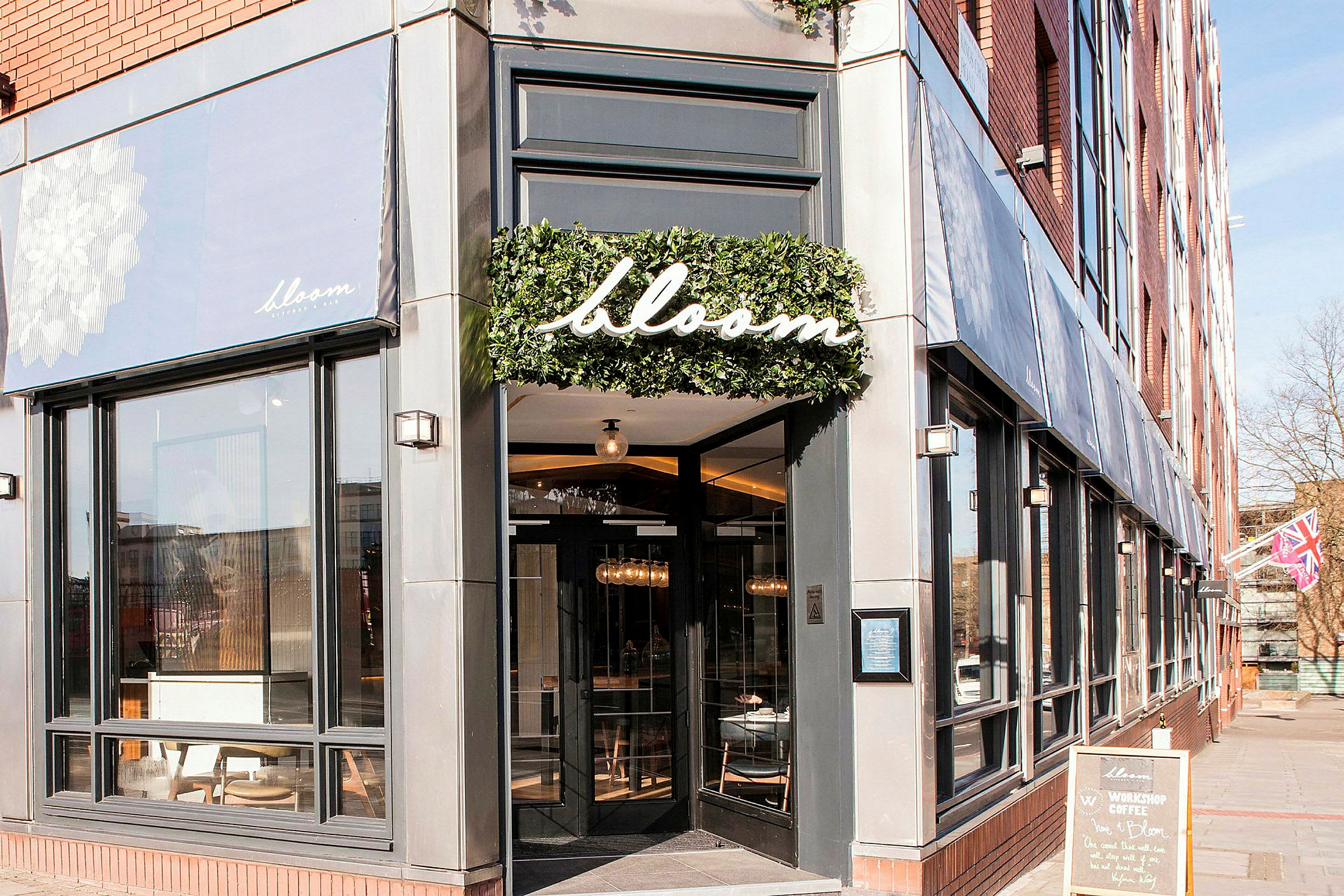 Inngangen til restauranten Bloom med store vinduer og et skilt med navnet omgitt av grønt. (AI-generert tekst)