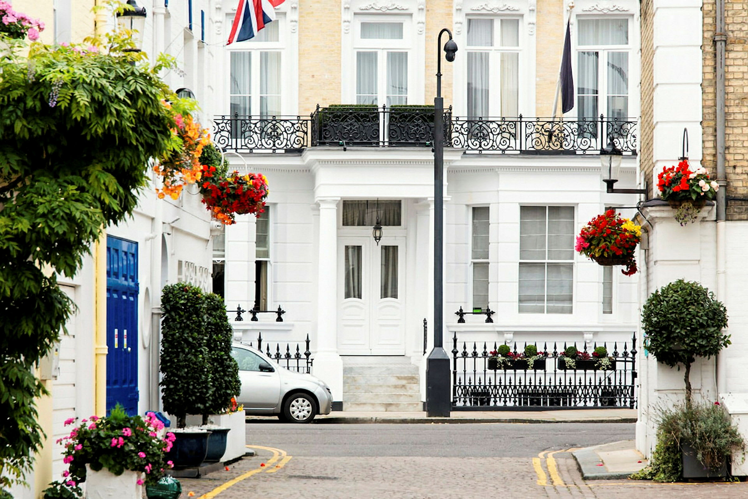 En hvit bygning med dekorative jernrekkverk, blomsterarrangementer og en bil parkert foran. Storbritannias flagg er synlig. (AI-generert tekst)