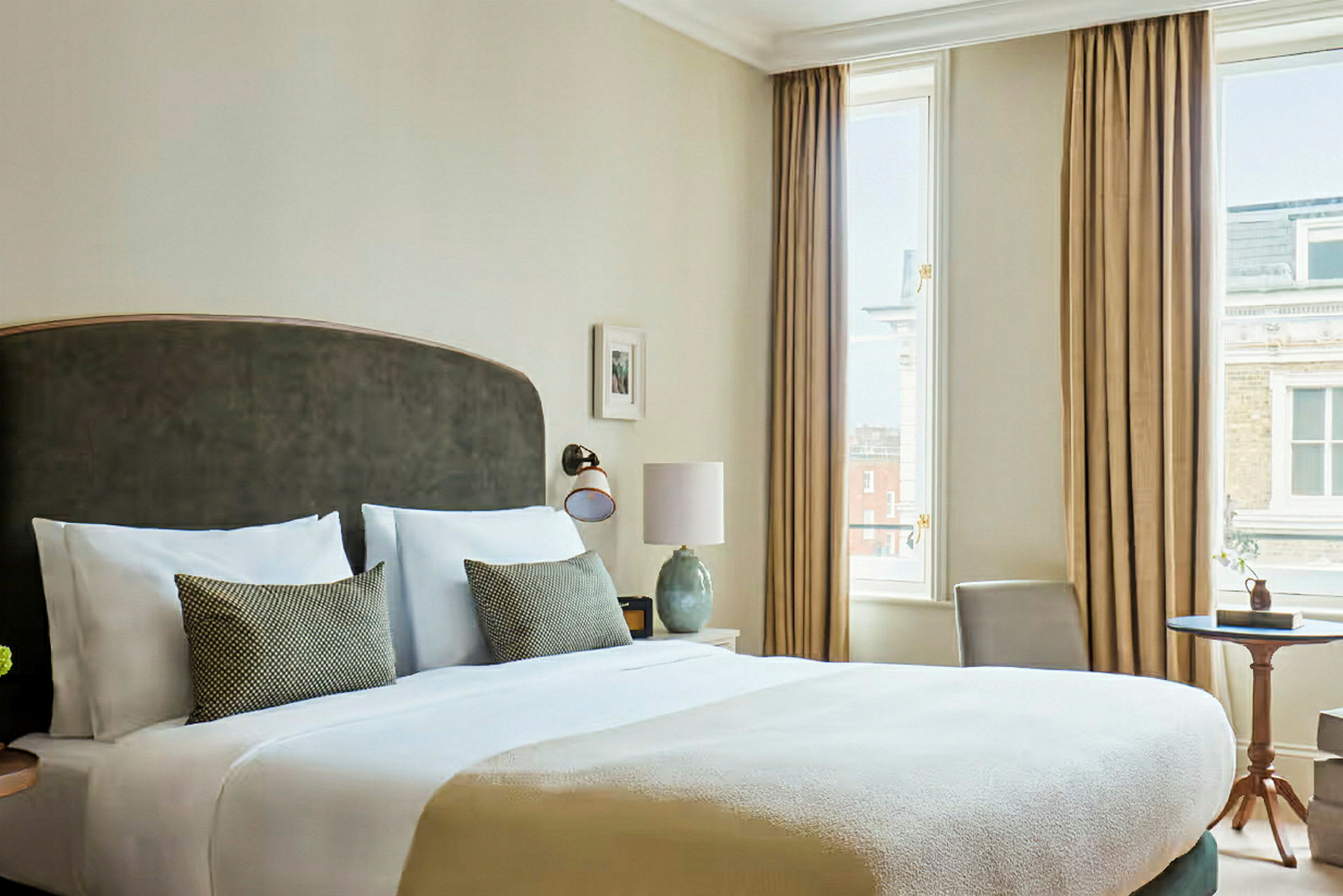 Et elegant hotellrom med en stor seng, to puter, et nattbord og store vinduer med gardiner. (AI-generert tekst)