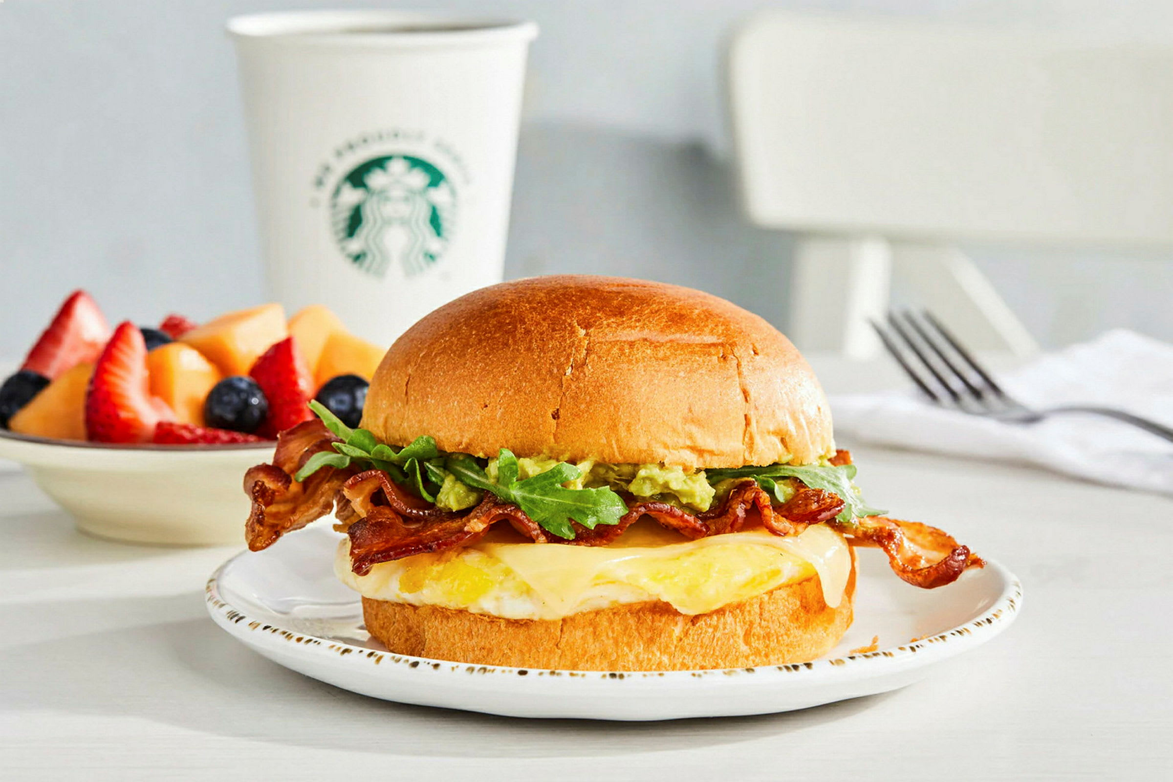 En tallerken med en sandwich som inneholder bacon, salat, ost og et stekt egg. I bakgrunnen en kopp med merke og en bolle med fersk frukt. (AI-generert tekst)