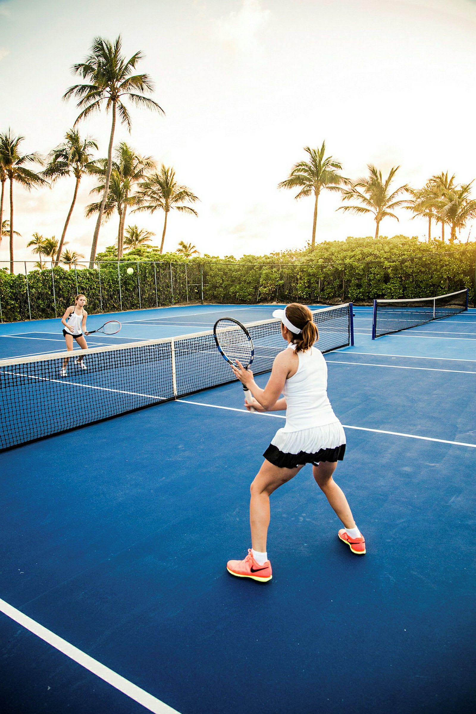 To personer spiller tennis på en utendørsbane omgitt av palmer. (AI-generert tekst)