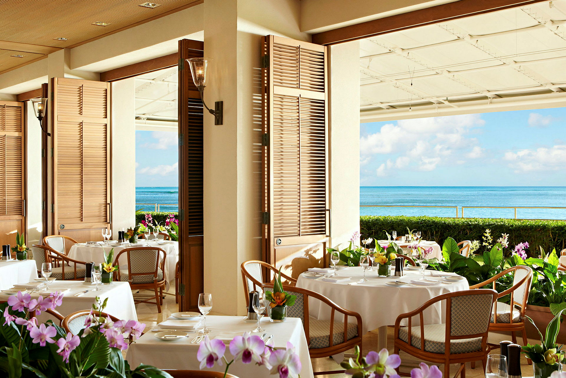 En restaurant med bord dekket med hvite duker og blomster. Bakgrunnen viser havutsikt med blå himmel. (AI-generert tekst)