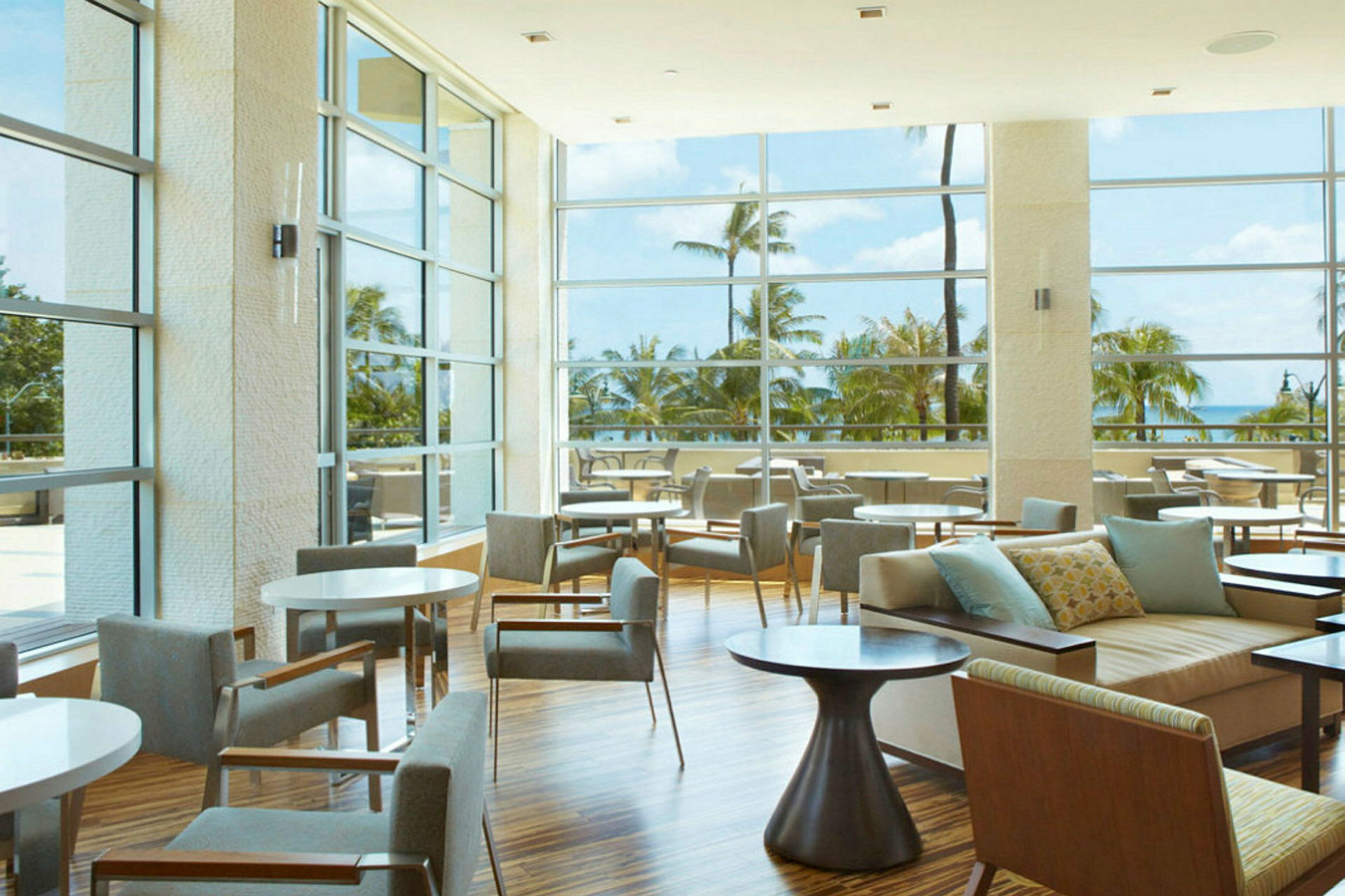 En hotellounge med store glassvinduer som gir utsikt mot palmer og havet. (AI-generert tekst)