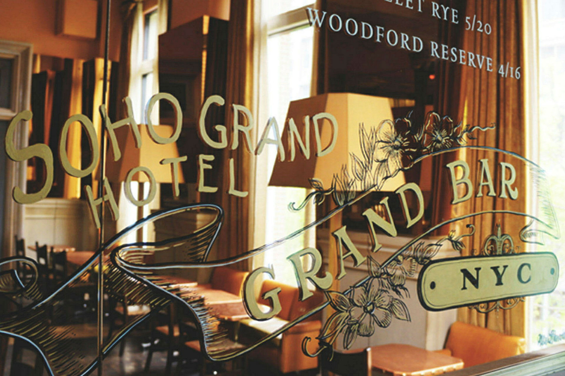 Et vindu med teksten 'Soho Grand Hotel' og 'Grand Bar NYC,' samt dekorasjoner. Bakgrunnen viser møbler og lamper. (AI-generert tekst)