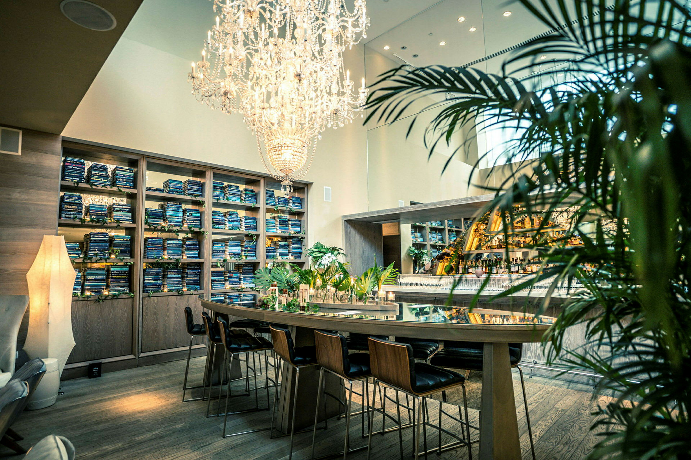 En elegant lokal med en lysekrone over et langbord med stoler, et barområde i bakgrunnen og hyller fylt med bøker. (AI-generert tekst)