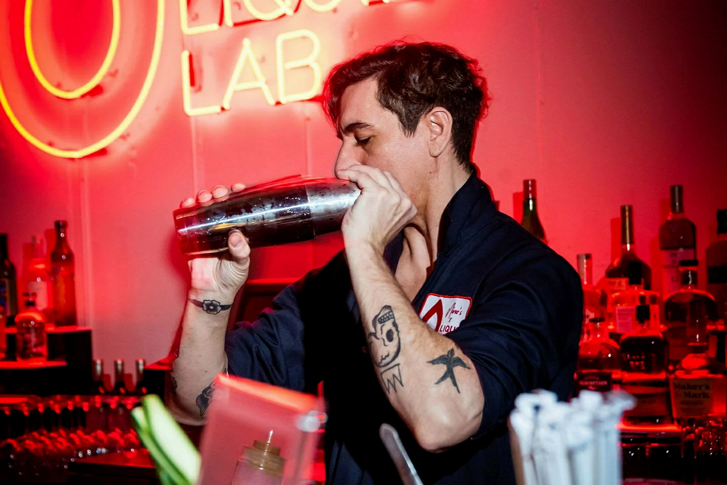 En person med tatoveringer holder en cocktailshaker i en bar med rød belysning. (AI-generert tekst)
