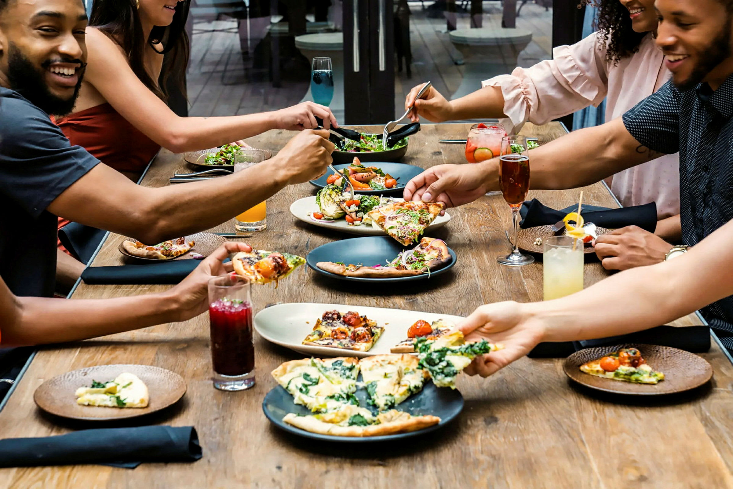 En gruppe mennesker spiser mat ved et bord. Det serveres pizza og andre retter, sammen med forskjellige drikker. (AI-generert tekst)