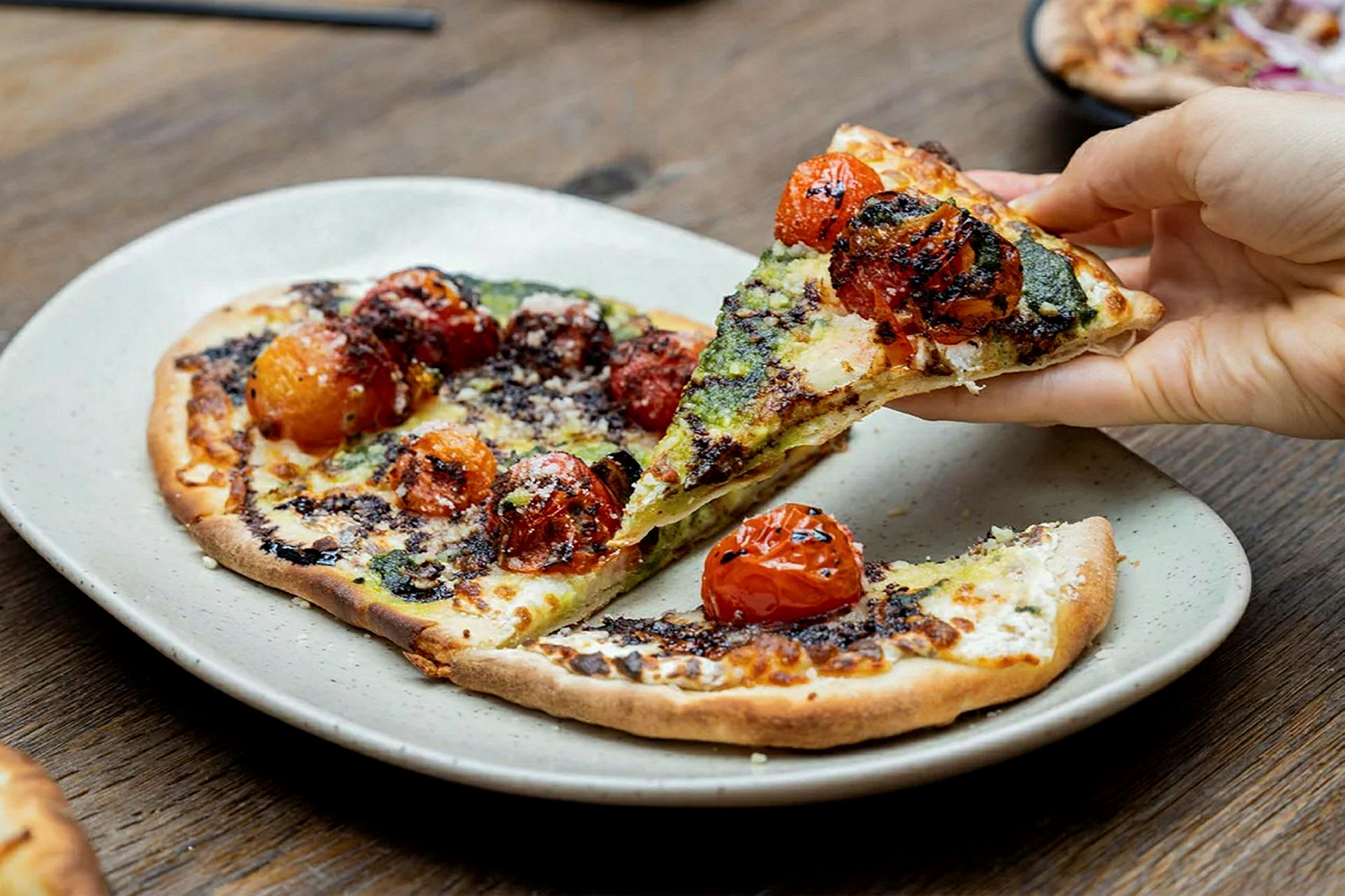 En tallerken med en pizza toppet med ristede tomater og pesto, hvor noen tar et stykke. (AI-generert tekst)