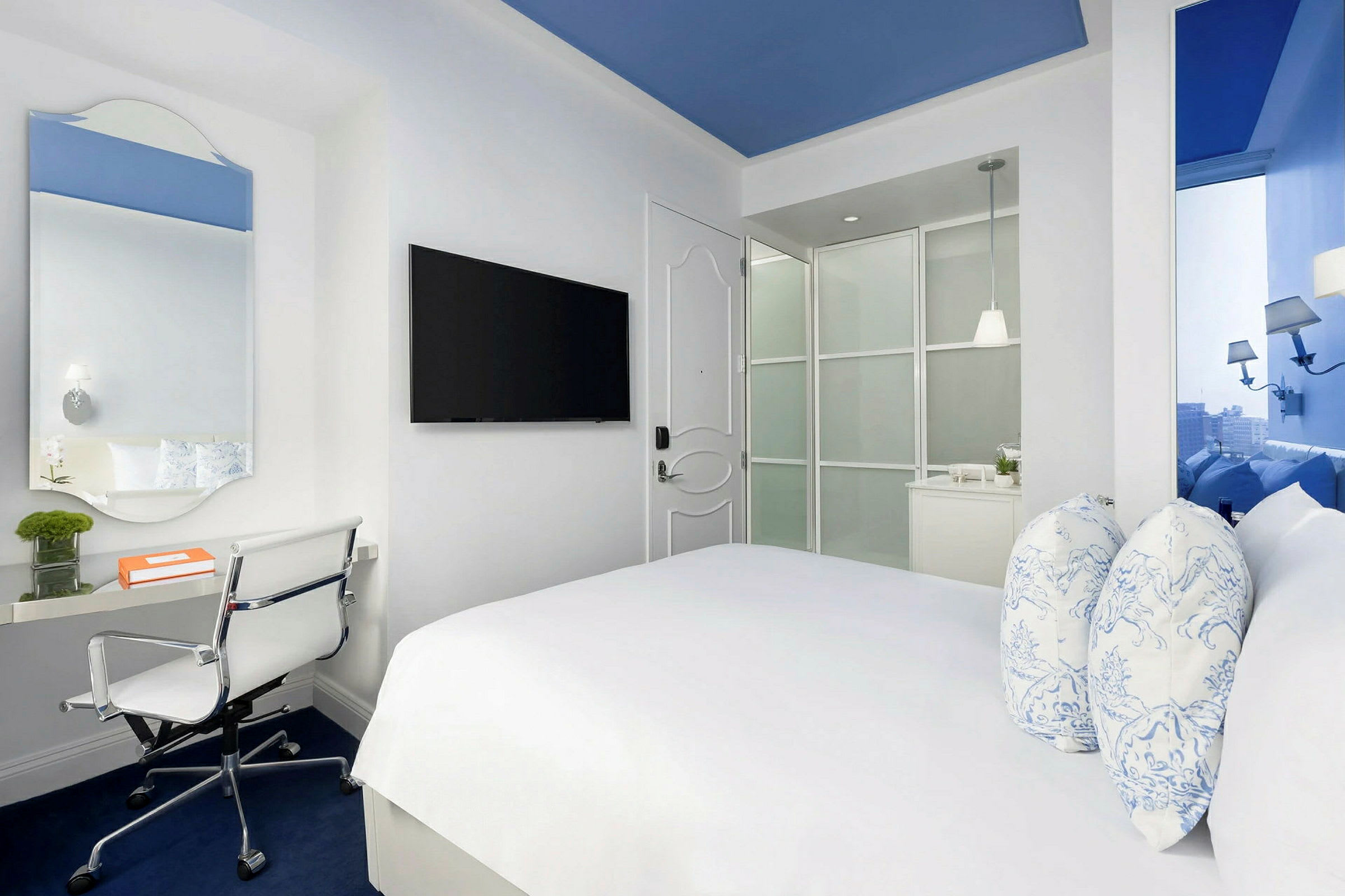 Et moderne hotellrom med en dobbeltseng, skrivebord og stol samt en veggmontert TV. Rommet er lyst med hvite vegger og blå detaljer. (AI-generert tekst)